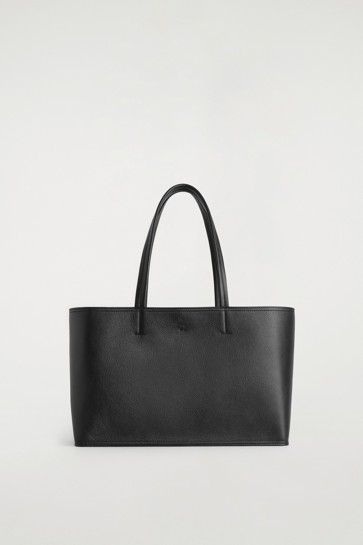 A black pebbled leather tote bag stands upright and faces forward, featuring a structured rectangular silhouette and two slim, rounded top handles. The bag displays a textured finish, clean stitched edges, and a flat base.