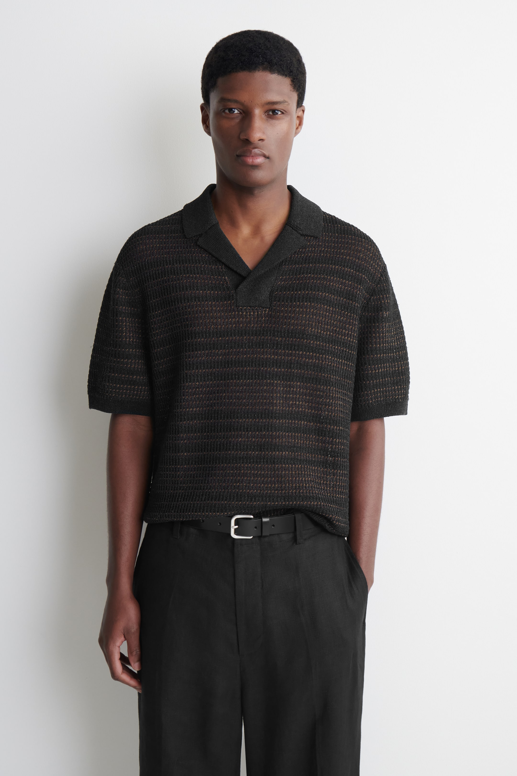 Agrandir l'image: Un polo noir à manches courtes est présenté de face, avec une construction en maille ajourée qui crée des bandes horizontales de différentes opacités. Il est conçu avec un col évasé et une patte de boutonnage en V.