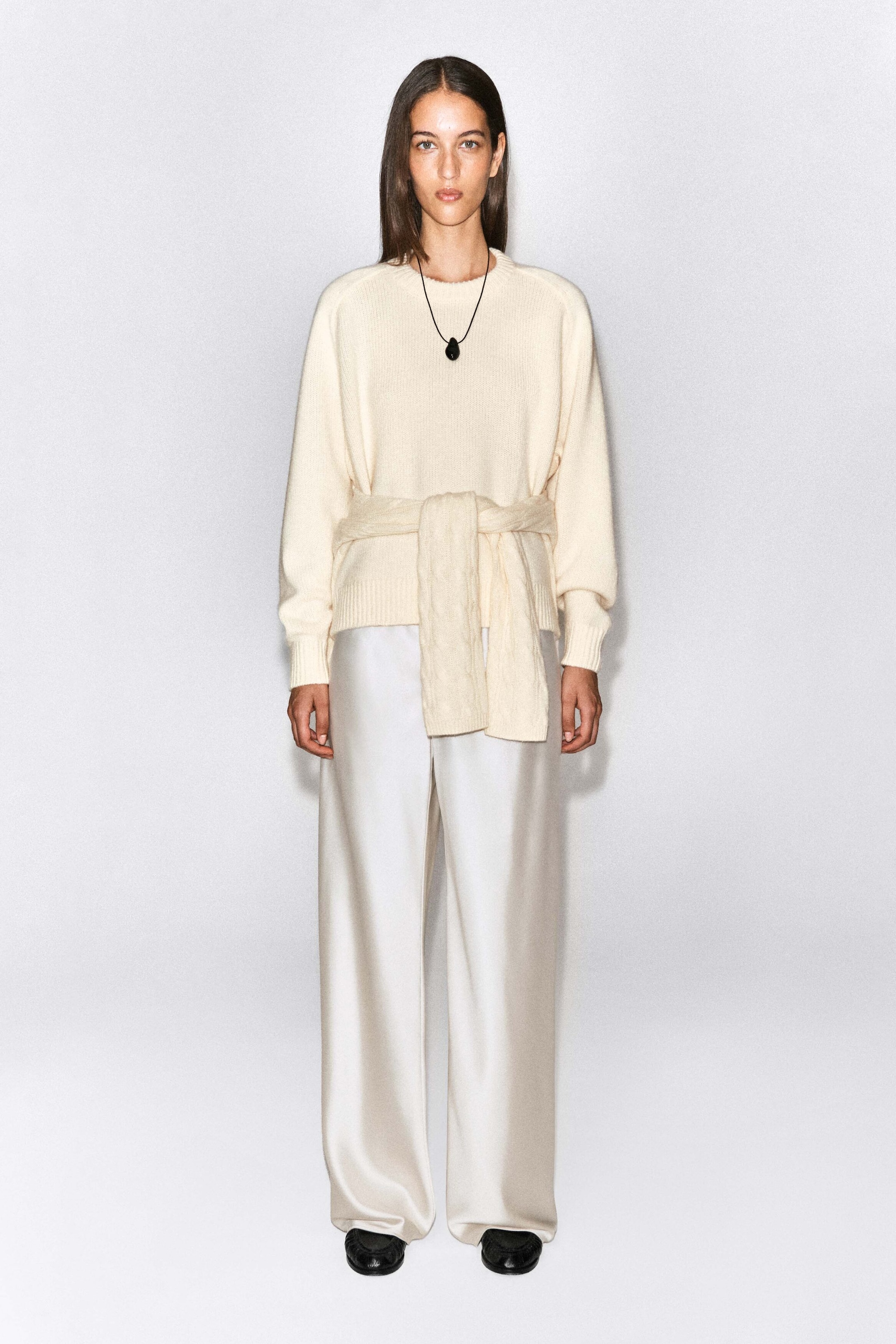 A rounded cashmere crew-neck sweater in cream is being worn by the model, featuring long sleeves, ribbed cuffs, and a relaxed silhouette. The sweater is styled with a matching knit piece tied around the waist and paired with wide-leg satin trousers. A rounded cashmere crew-neck sweater in cream is being worn by the model, featuring long sleeves, ribbed cuffs, and a relaxed silhouette. The sweater is styled with a matching knit piece tied around the waist and paired with wide-leg satin trousers.