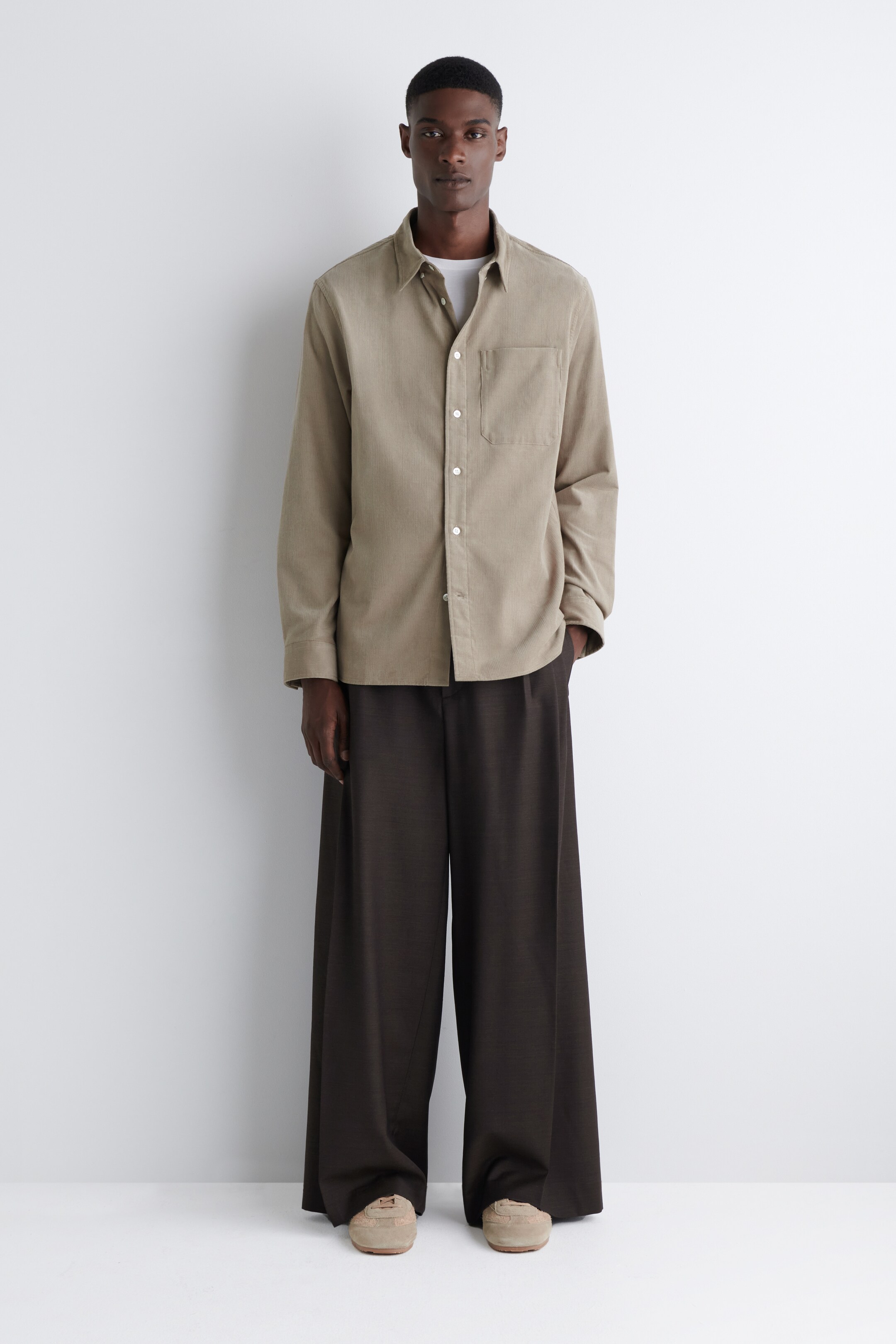 View larger image: A light brown ribbed long-sleeved button-up shirt, featuring a single chest pocket and white buttons, is being presented front-on, layered over a white crew-neck t-shirt. Dark brown wide-leg trousers are being styled with the shirt, and light beige low-top sneakers are completing the look.
