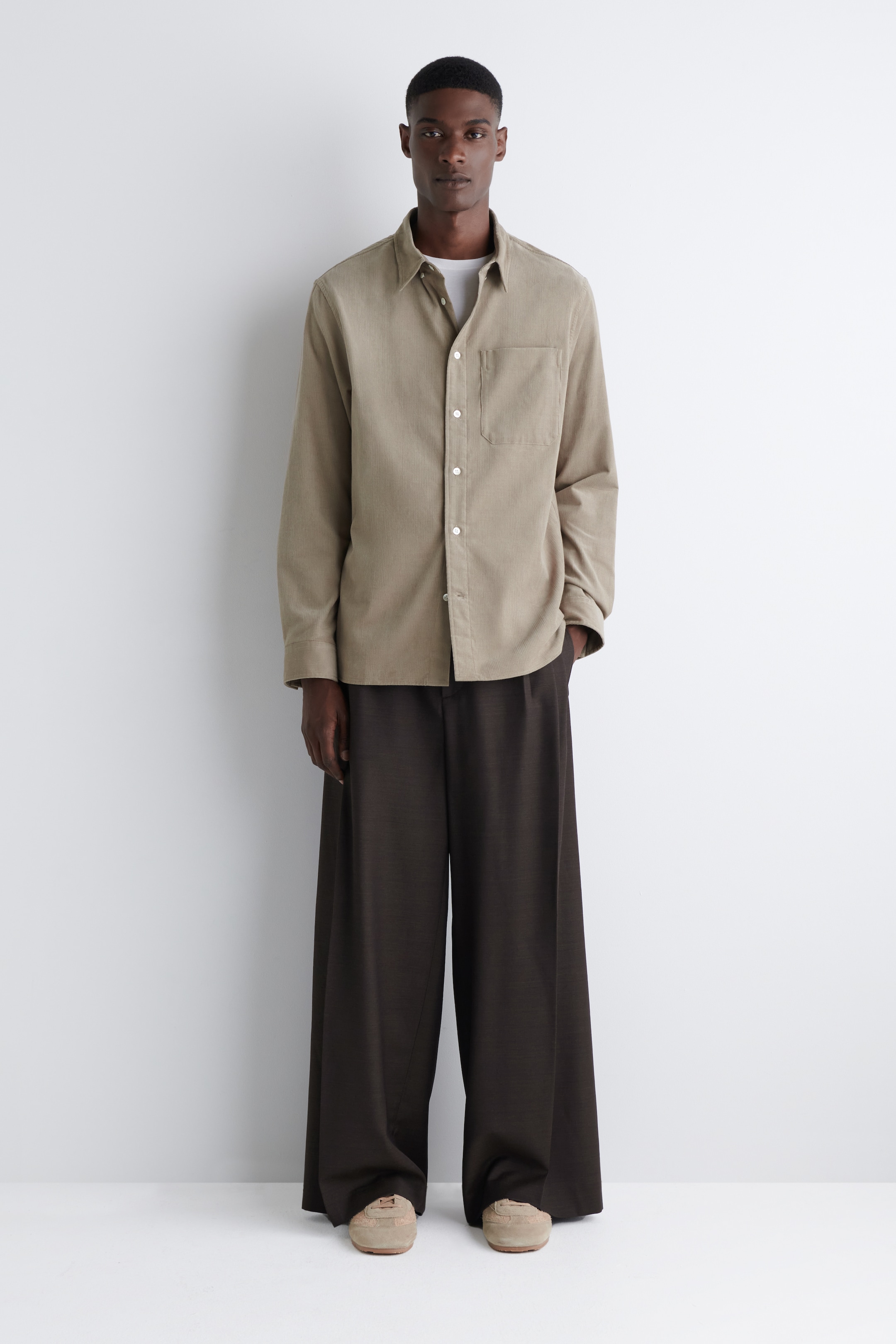 Agrandir l'image: Une chemise à manches longues boutonnée de couleur brun clair côtelée, avec une poche poitrine unique et des boutons blancs, est présentée de face, superposée à un t-shirt à col rond blanc. Un pantalon large de couleur brun foncé est assorti à la chemise, et des baskets basses de couleur beige clair complètent le look.