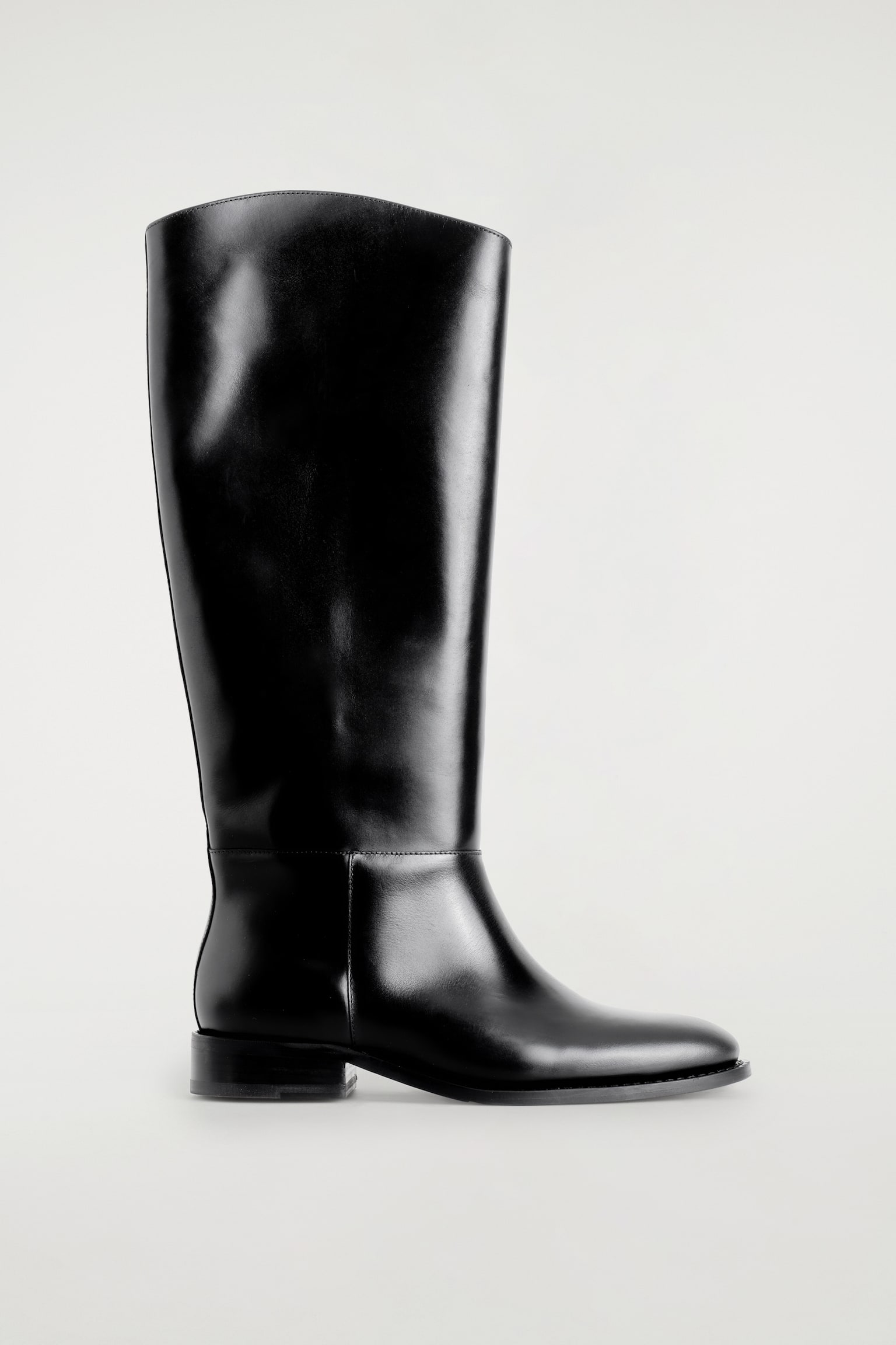 Une botte dâÃ©quitation noire en cuir, montant jusquâau genou, est prÃ©sentÃ©e de profil, mettant en valeur une finition polie, une ligne supÃ©rieure galbÃ©e et sculptÃ©e, un bout arrondi et un petit talon carrÃ©.