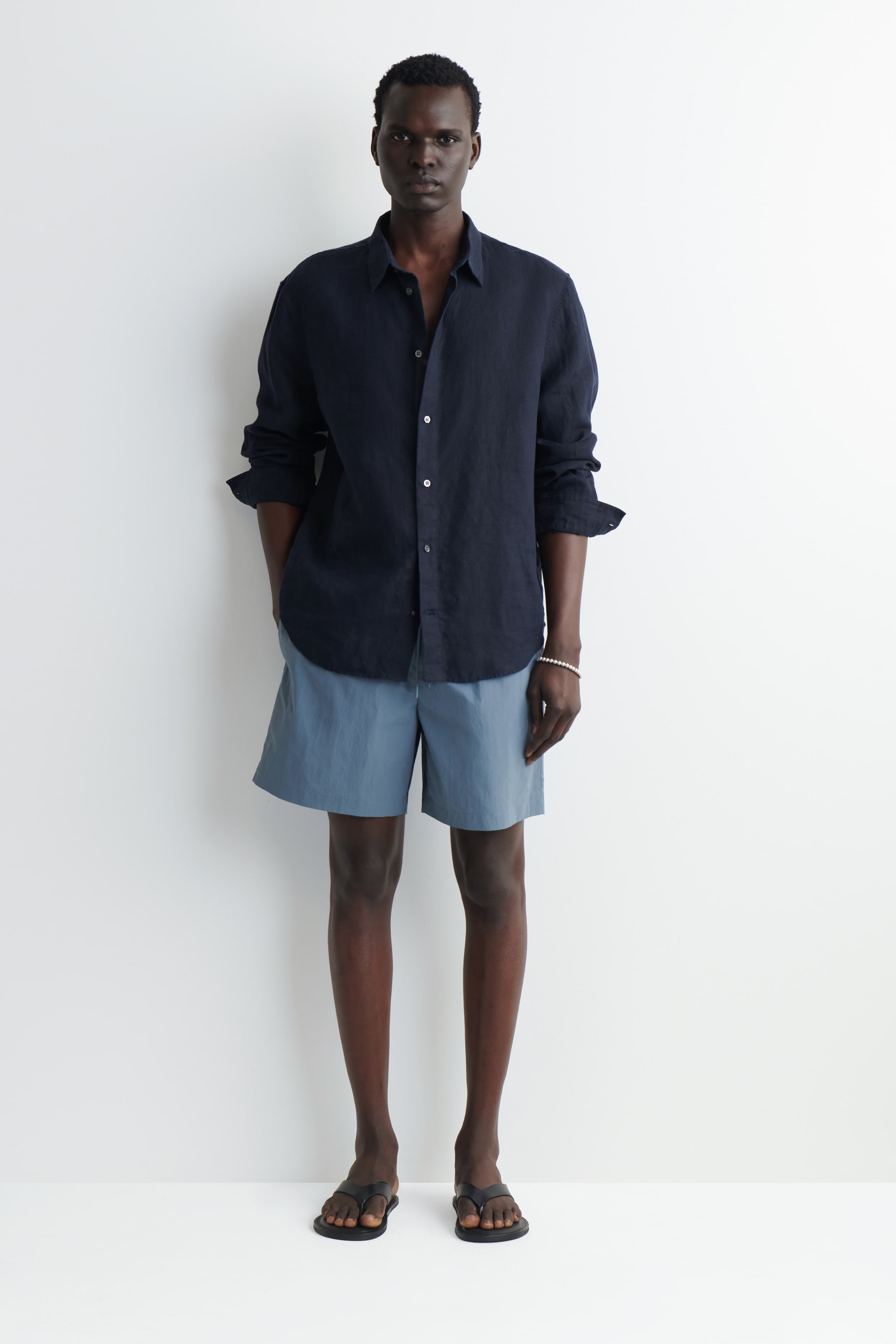 Visualizza immagine più grande: Una camicia in lino blu navy con bottoni è indossata fuori dai pantaloni con le maniche arrotolate, abbinata a shorts blu ardesia con coulisse visibile e sandali infradito neri.