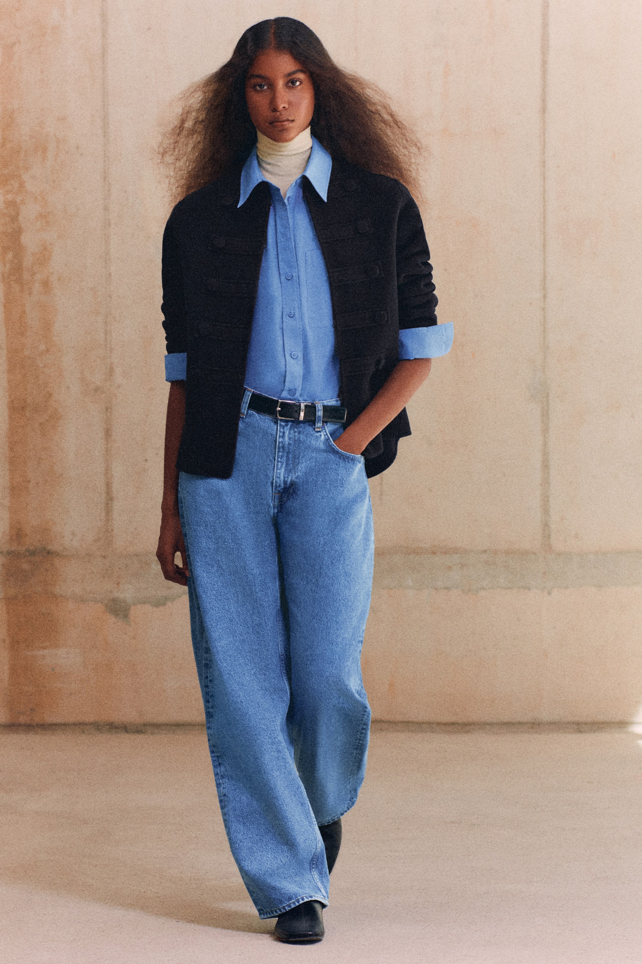Größeres Bild anzeigen: Ein Model trägt eine schwarze Uniformjacke aus doppelseitiger Wolle und hellblau gewaschene Jeans mit gebogenem Bein und aufgesetztem Einsatz, von vorne gesehen. Die strukturierte Jacke verfügt über horizontale, militärisch inspirierte Banddetails, während die hoch geschnittene, locker sitzende Jeans eine markante, gebogene Beinsilhouette zeigt und mit einem schwarzen Gürtel gestylt ist.