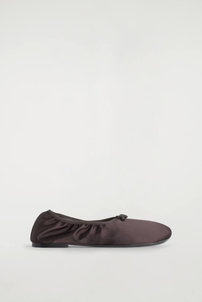 Ein einzelner Ballerina wird in einem dunkelbraunen Satin-Stoff präsentiert, mit einem elastischen Einstieg, der mit Ripsband eingefasst ist und einem kleinen Schleifendetail am Vorderblatt. Der Schuh hat eine abgerundete Spitze und einen niedrigen, flachen Absatz. - 1
