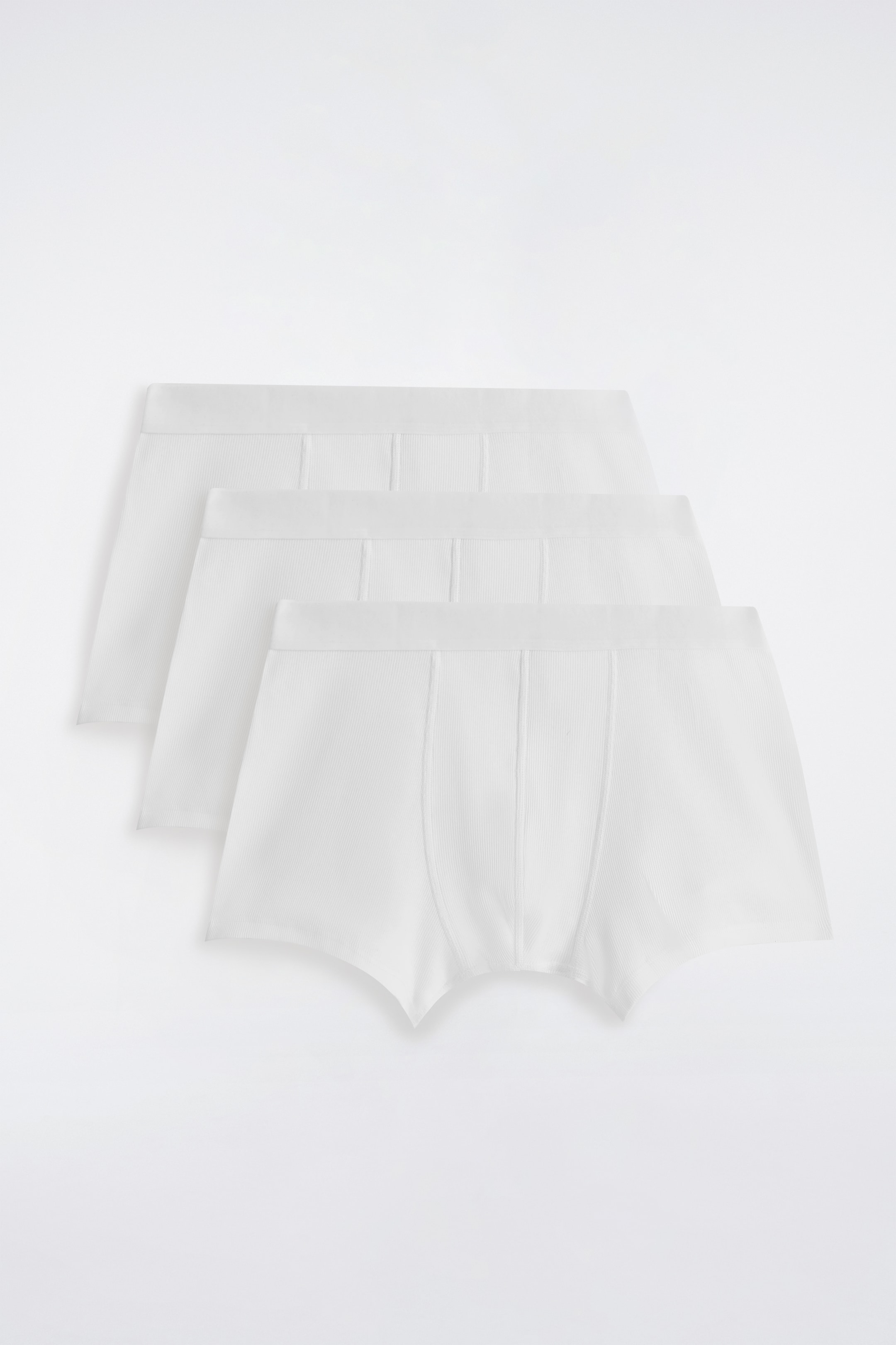 Grotere afbeelding bekijken: Drie witte ribgebreide boxershorts zijn gestapeld en overlappen elkaar, gepresenteerd met hun voorkanten naar voren gericht. Elke slip is voorzien van een elastische tailleband, zichtbare verticale stiksels op de voorzak en een karakteristieke ribstructuur over de stof.