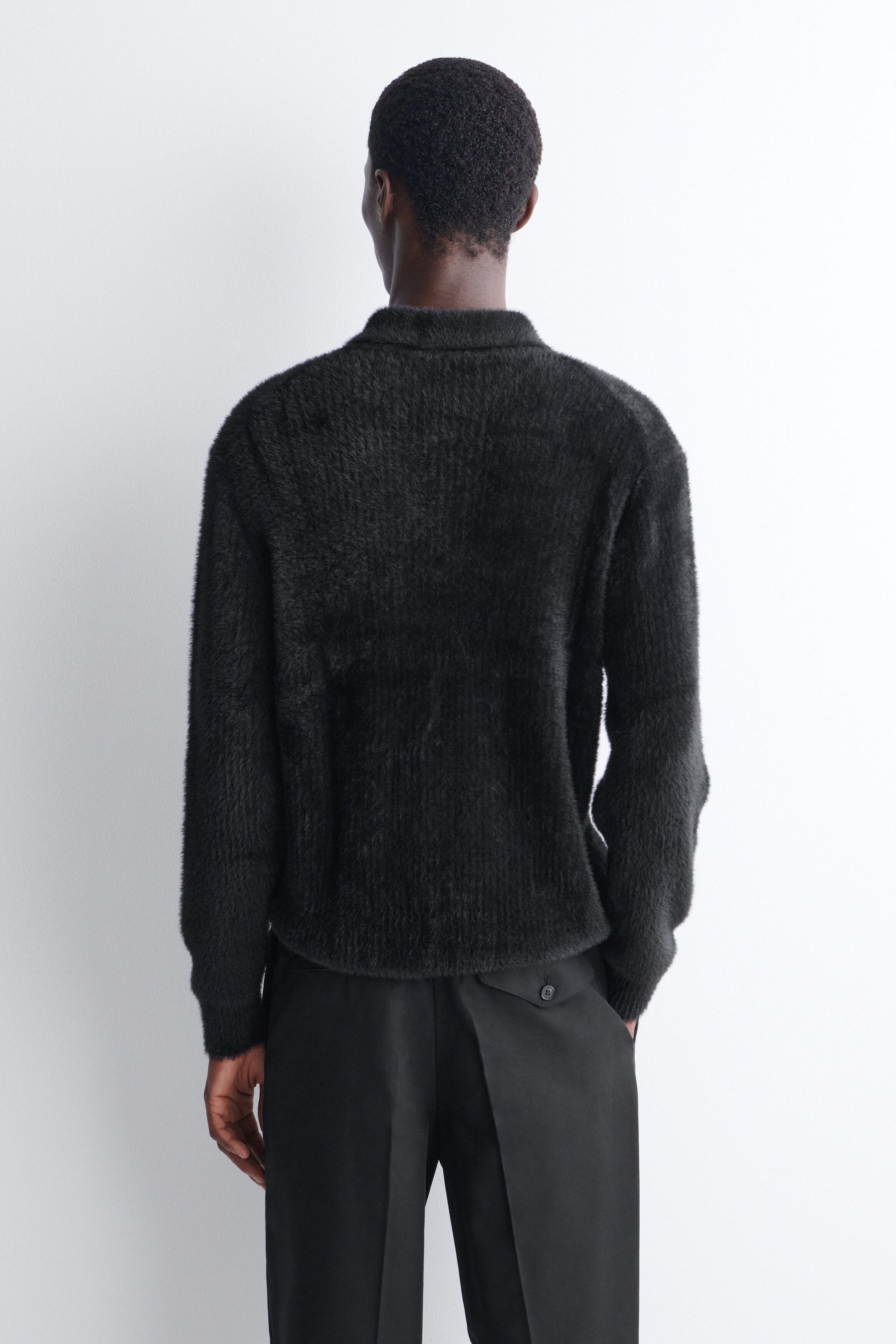 A black knitted polo shirt with a highly textured, brushed finish is being worn by the model, shown from the back. The long-sleeved shirt features a ribbed collar and cuffs and is styled with black trousers. A black knitted polo shirt with a highly textured, brushed finish is being worn by the model, shown from the back. The long-sleeved shirt features a ribbed collar and cuffs and is styled with black trousers.