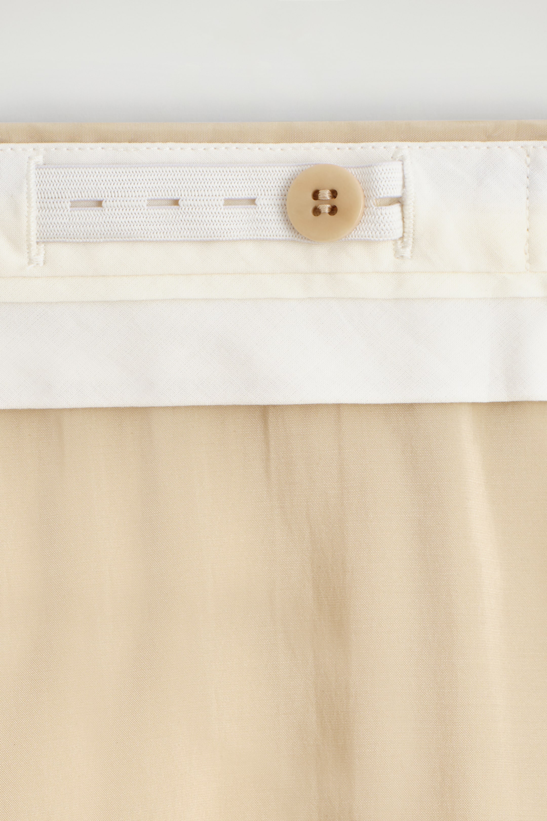 Zobraziť väčší obrázok: FLUIDNÉ PLISOVANÉ CULOTTES zobrazujú detailný pohľad na ich vnútorný nastaviteľný pás, ktorý obsahuje bielu elastickú pásku s viacerými dierkami na gombíky a svetlohnedý gombík na úpravu pásu, zatiaľ čo ich fluidná, svetlo béžová látka sa jemne skladá a ukazuje jemné záhyby nižšie.
