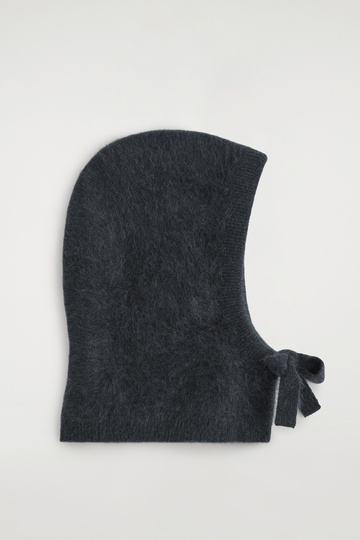A dark gray brushed cashmere hood is shown in side profile, highlighting its soft, fuzzy texture and ribbed face opening. A matching knitted tie forms a neat bow at the front of the neck.