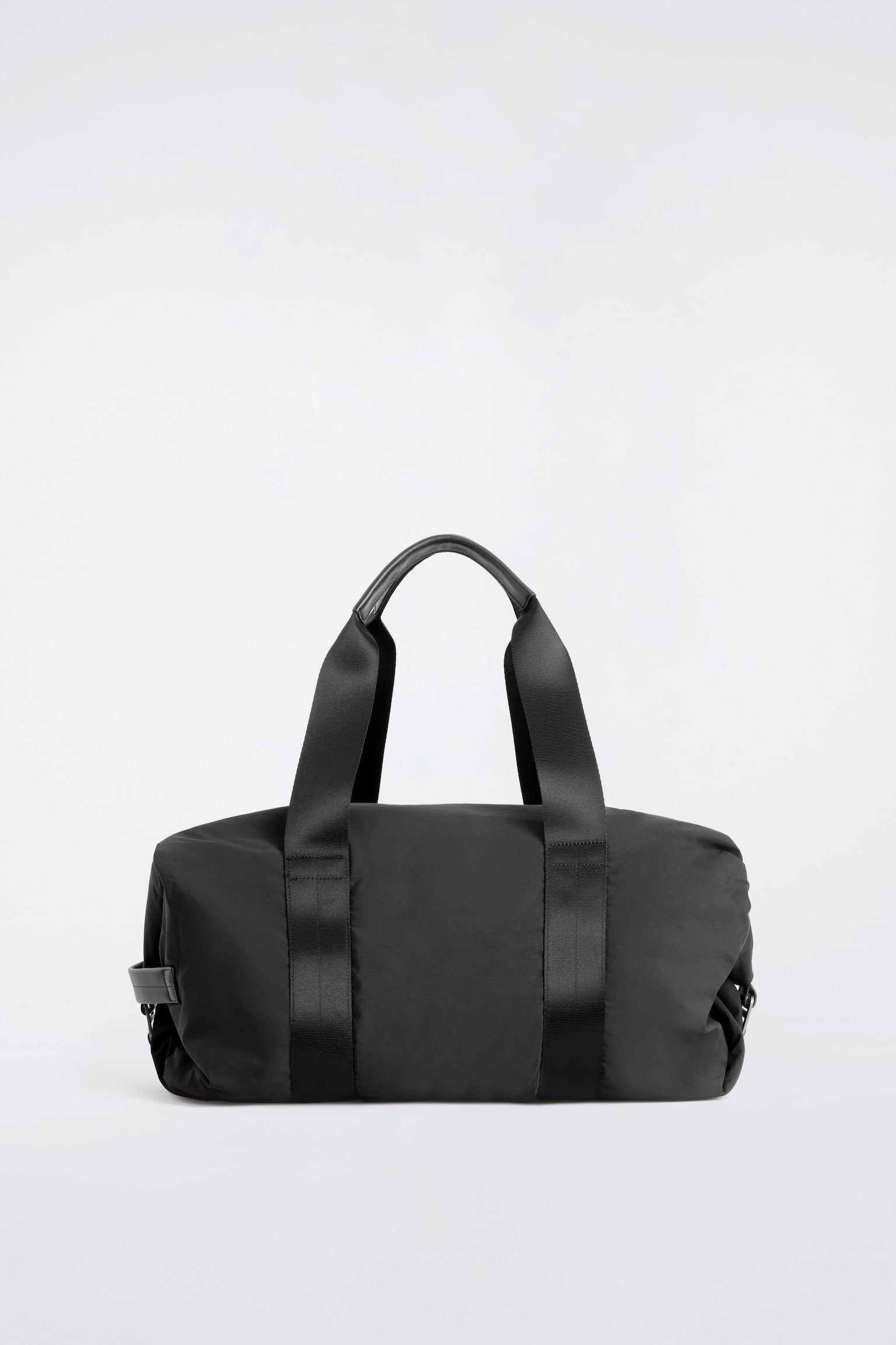 Die schwarze Nylon-Duffle-Bag ist in einer frontalen Ansicht platziert und zeigt ihren weich strukturierten, zylindrischen Korpus. Zwei breite Webbing-Riemen verlaufen vertikal entlang der Seiten und fÃ¼hren nach oben, um Tragegriffe zu bilden, die in der Mitte ein glattes Ledergriffdetail aufweisen.