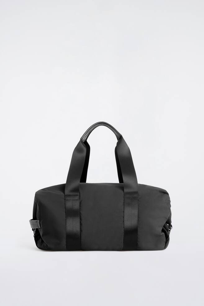 Le sac de voyage noir en nylon est prÃ©sentÃ© de face, mettant en valeur sa silhouette cylindrique Ã  structure souple. Deux larges sangles en sangle descendent verticalement le long des cÃ´tÃ©s, puis se prolongent vers le haut pour former des poignÃ©es supÃ©rieures, agrÃ©mentÃ©es dâun dÃ©tail de prise en main en cuir lisse au centre. - 1