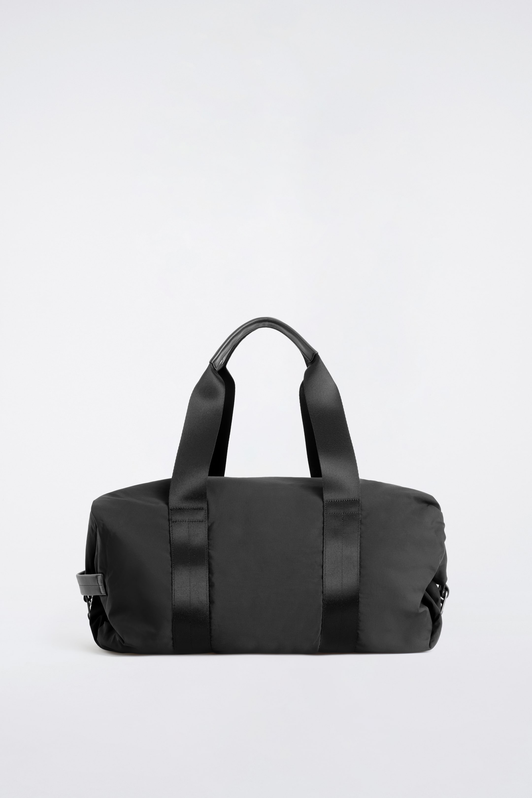 View larger image: The black nylon duffle bag is shown from the front, highlighting its softly structured cylindrical silhouette. Two wide webbing straps run vertically down the sides and extend upward to form top handles, each featuring a smooth leather grip detail at the centre.
