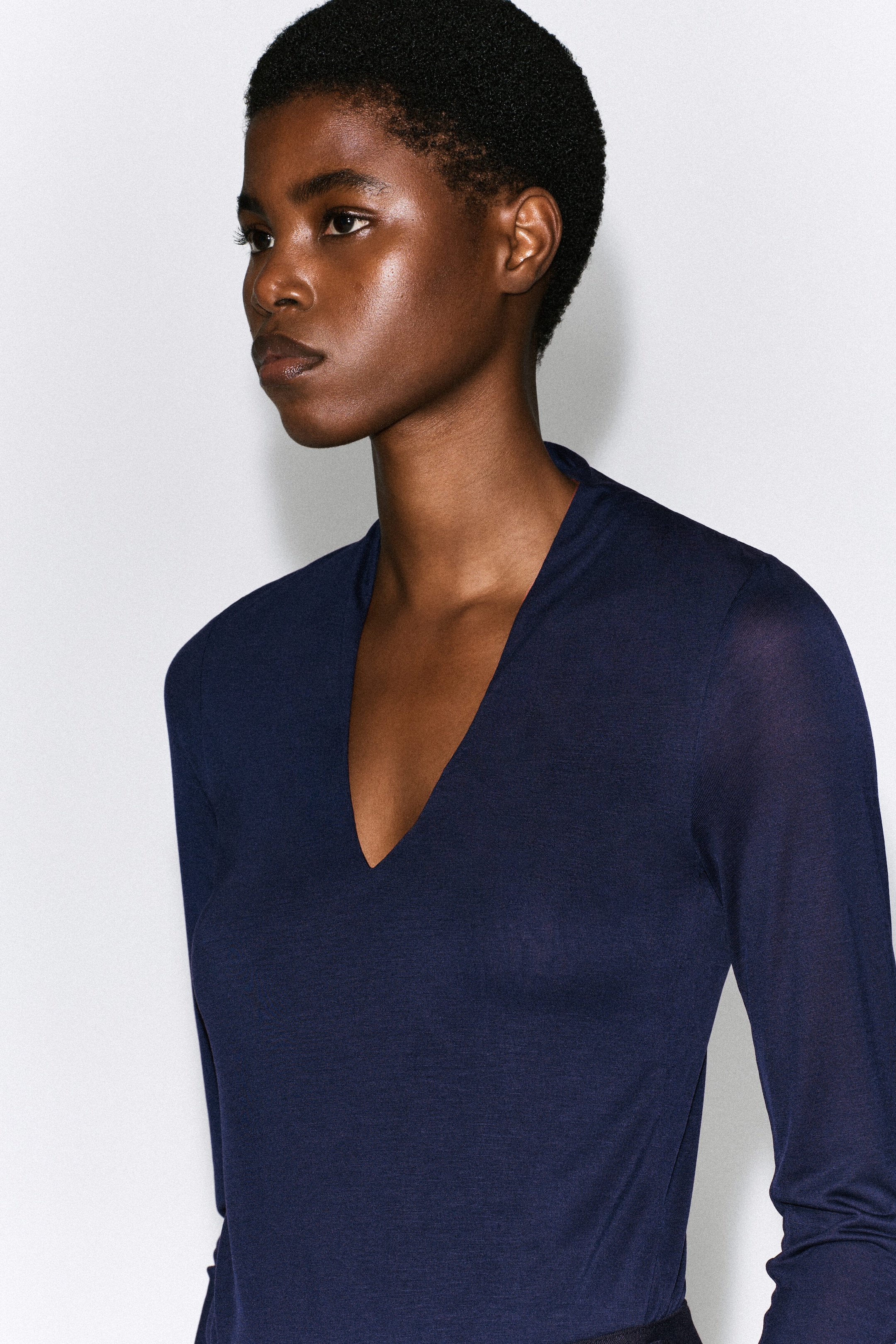 The model is wearing a navy blue, long-sleeved top with a deep V-neckline. The garment features a slim-fit silhouette and is made from a lightweight, fine-knit jersey fabric. The model is wearing a navy blue, long-sleeved top with a deep V-neckline. The garment features a slim-fit silhouette and is made from a lightweight, fine-knit jersey fabric.