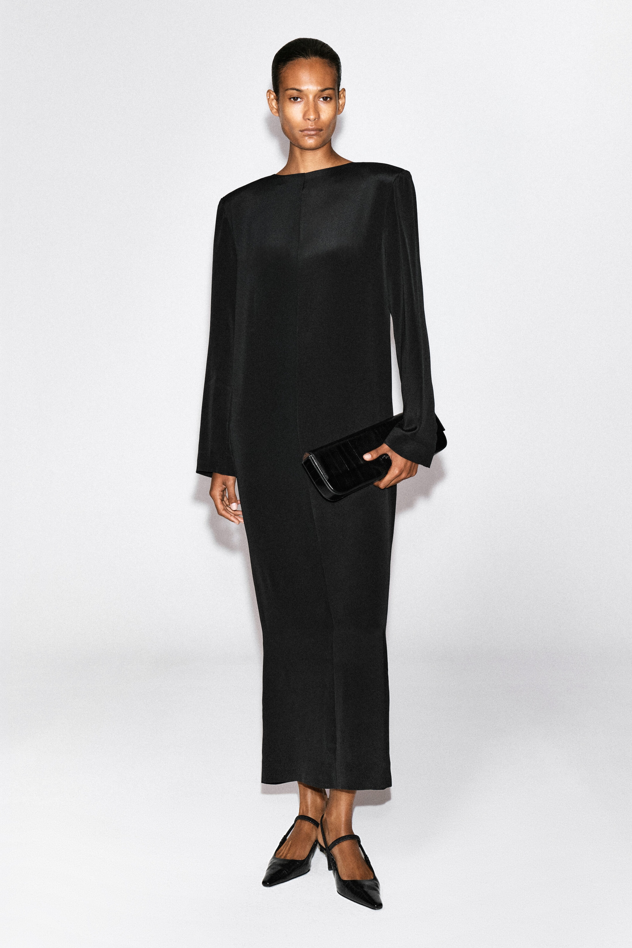 Ein schwarzes Maxikleid mit langen Ärmeln, strukturierten Schultern und einem hochgeschlossenen Rundhalsausschnitt wird vom Model getragen. Das Model hält eine schwarze Leder-Clutch und trägt schwarze Pumps mit spitzer Zehenpartie. Ein schwarzes Maxikleid mit langen Ärmeln, strukturierten Schultern und einem hochgeschlossenen Rundhalsausschnitt wird vom Model getragen. Das Model hält eine schwarze Leder-Clutch und trägt schwarze Pumps mit spitzer Zehenpartie.
