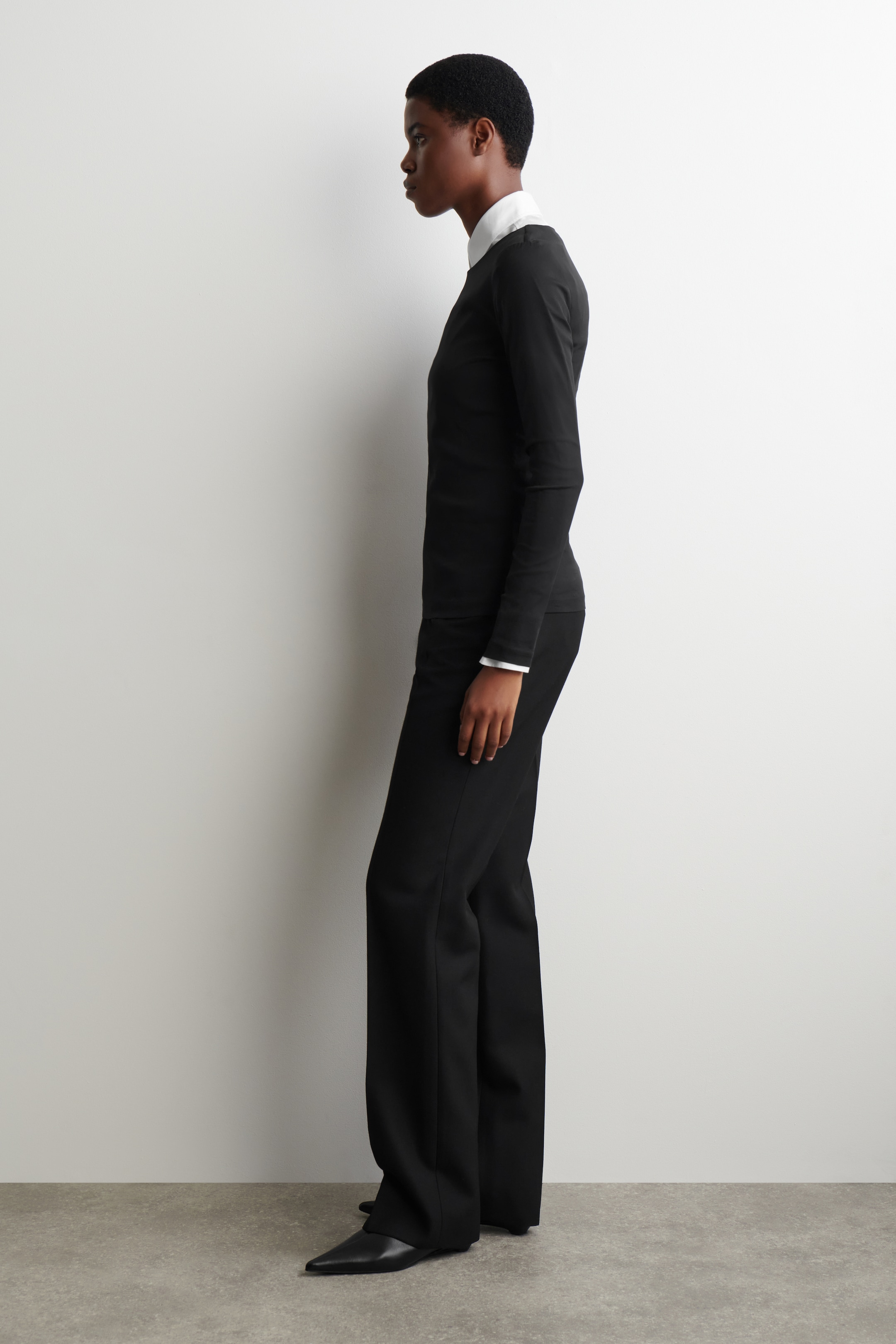 Grösseres Bild ansehen: Ein schwarzes Slim-Fit-Oberteil mit langen Ärmeln wird vom Model getragen. Das Oberteil wird als Basisschicht gestylt, wobei ein weißer Hemdkragen über dem Ausschnitt sichtbar ist und weiße Hemdmanschetten aus den Ärmeln hervorragen.