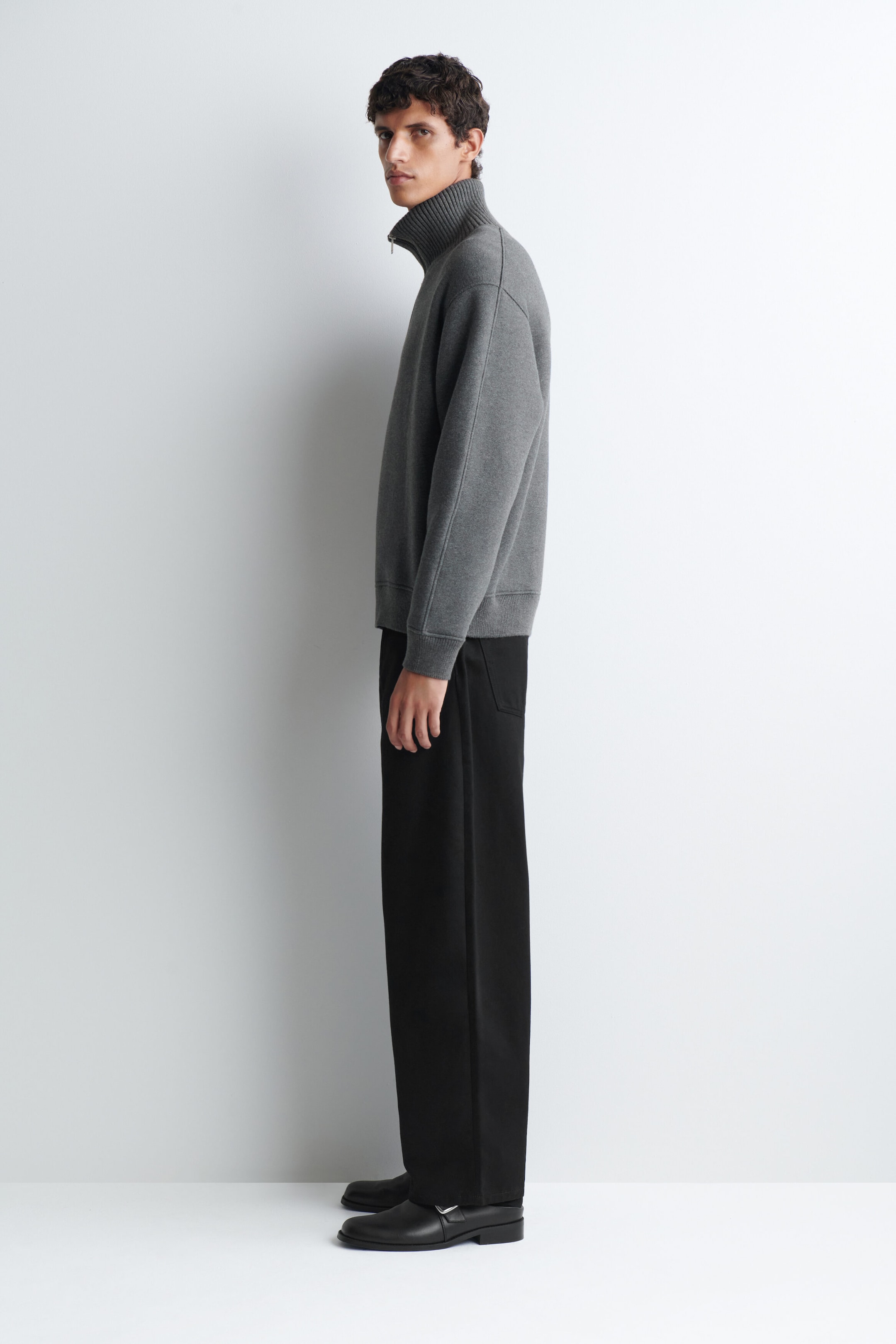 The model is wearing a grey cotton jersey zip-up jumper featuring a high ribbed collar, ribbed cuffs, and a ribbed hem. The jumper is styled with black wide-leg trousers and black shoes, as the model stands in side profile. The model is wearing a grey cotton jersey zip-up jumper featuring a high ribbed collar, ribbed cuffs, and a ribbed hem. The jumper is styled with black wide-leg trousers and black shoes, as the model stands in side profile.