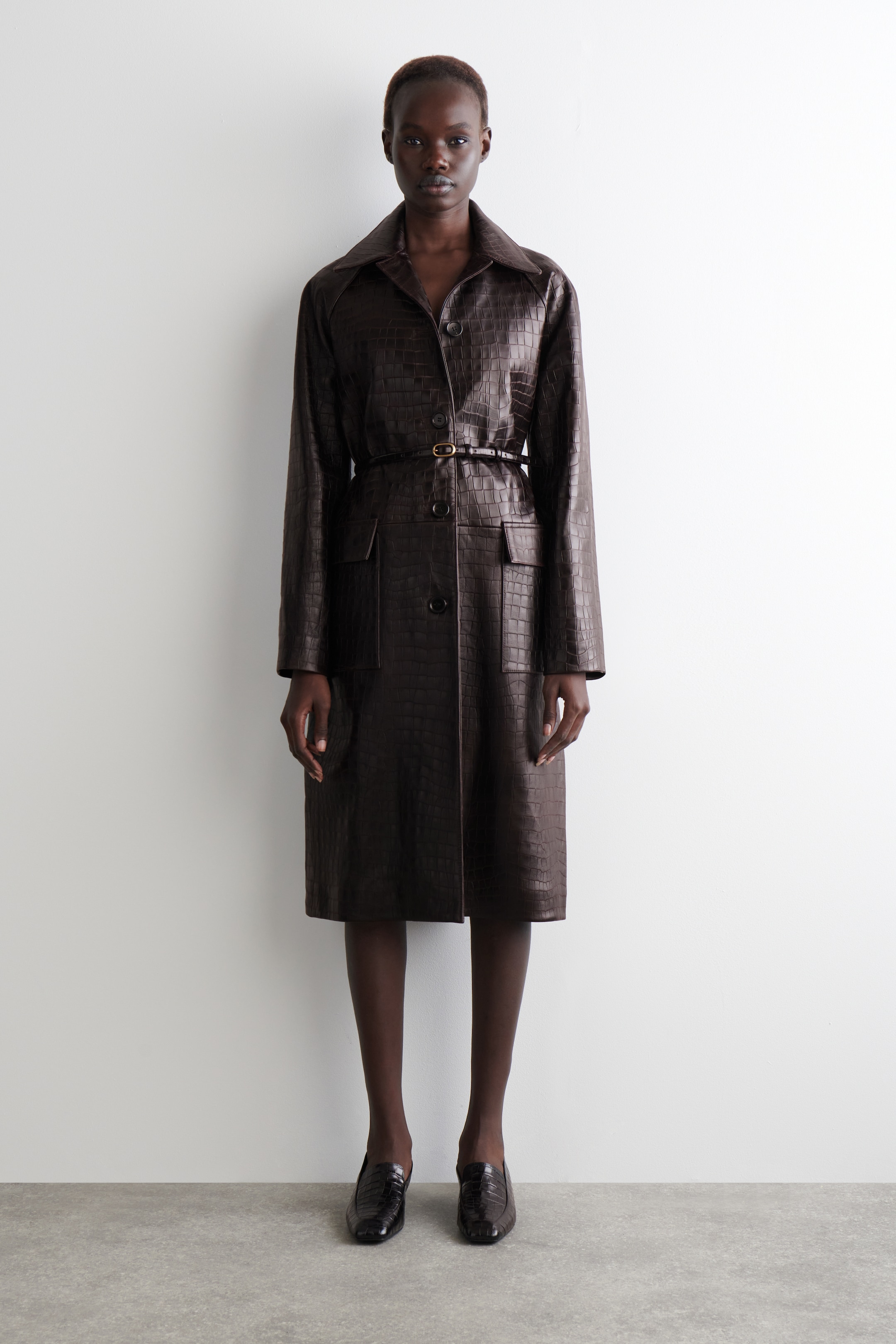 Agrandir l'image: Un manteau long brun foncé, embossé de motif crocodile, est exposé de face, en vue pleine longueur, avec un large col pointu, une fermeture à boutons sur le devant et deux grandes poches plaquées. Une fine ceinture assortie avec une petite boucle serre la taille.