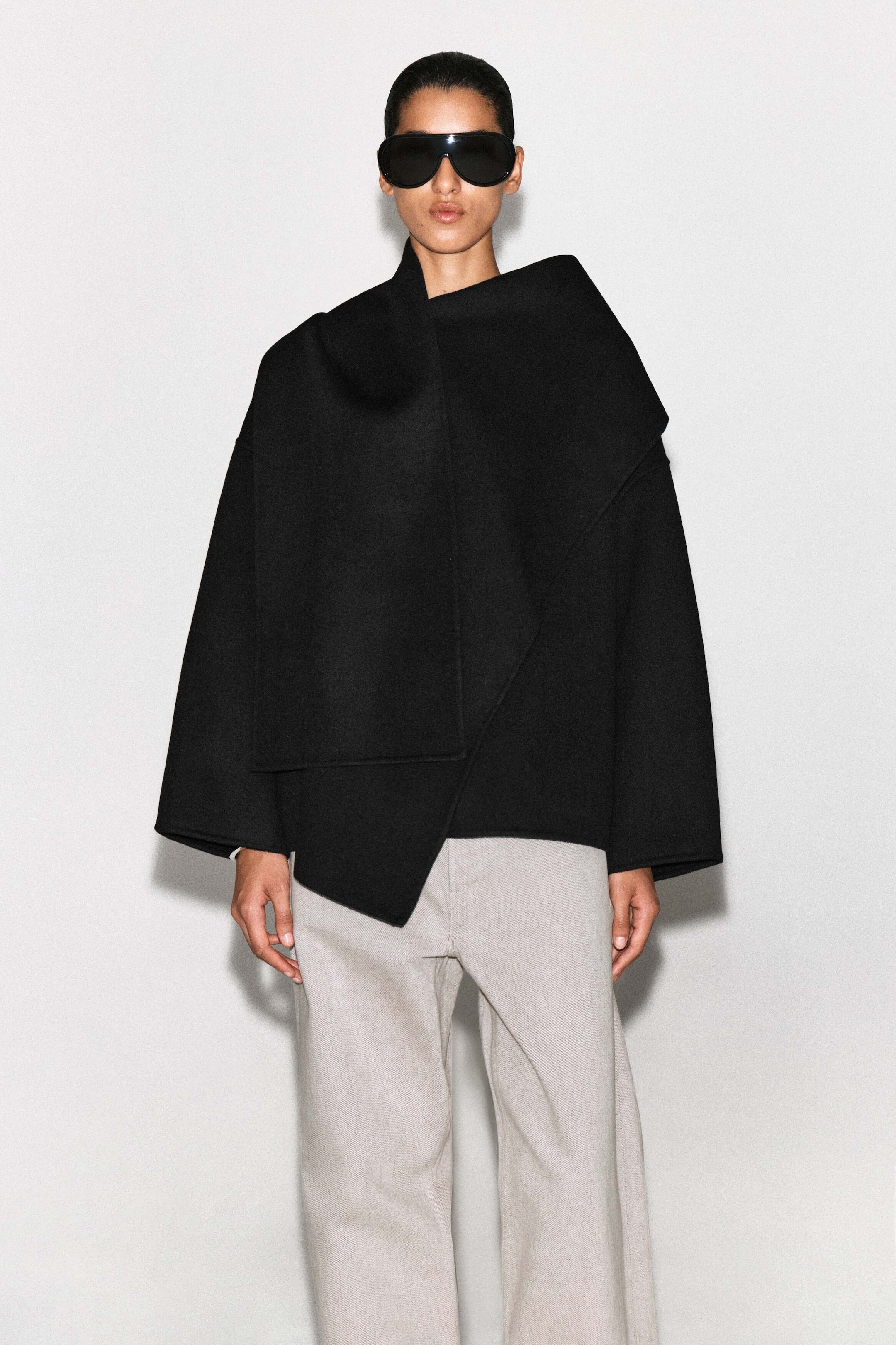A black double-faced wool-blend scarf jacket is shown from the front, featuring an oversized silhouette with an integrated scarf draped across the chest and shoulder. The jacket has wide, relaxed sleeves and an asymmetrical hemline, styled over light gray trousers.