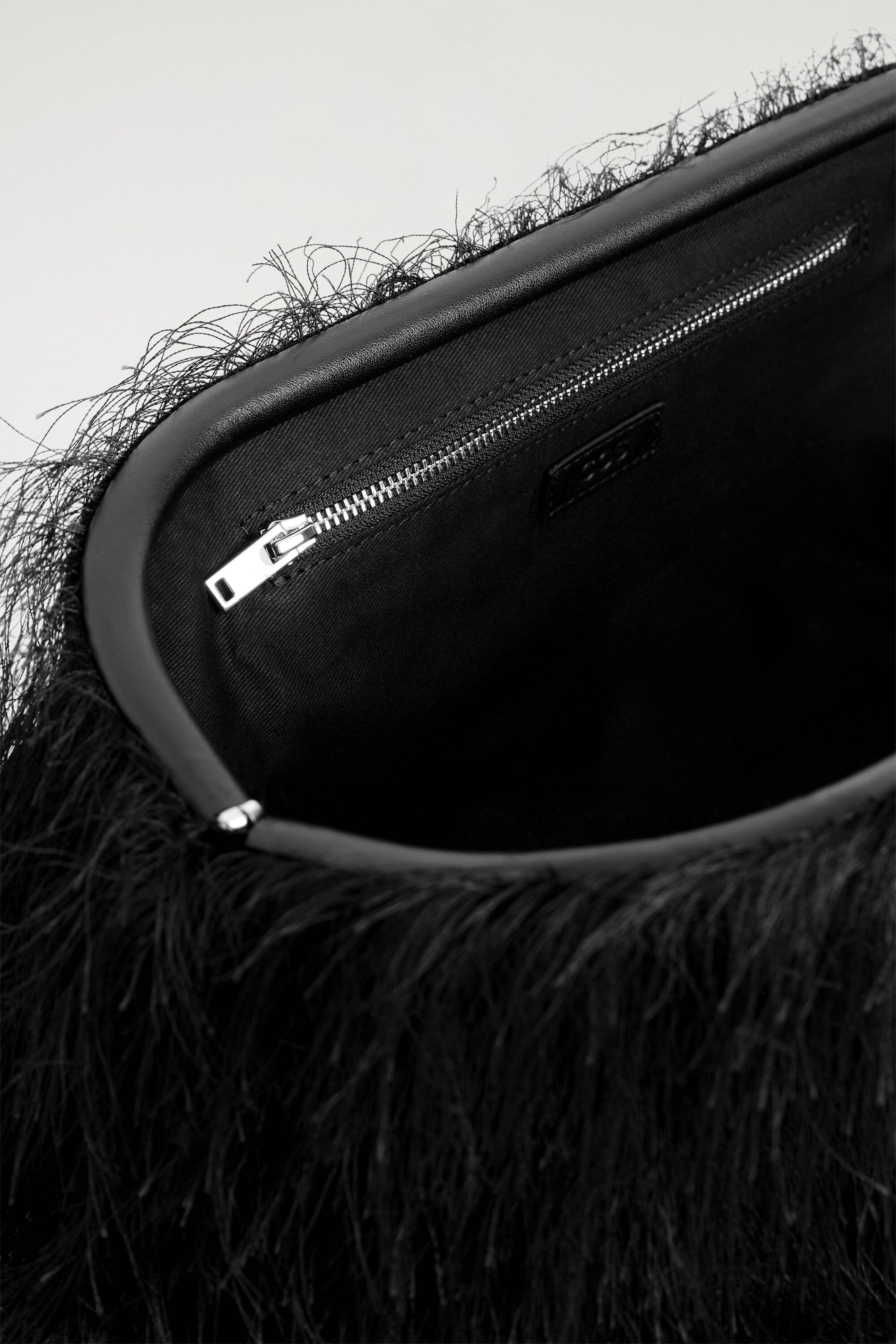 A black fringed clutch bag is showing its open interior, which is lined with black fabric and features a silver-toned zipped pocket. The smooth, rounded black leather frame is highlighting the dense, fine black fringe covering the bag's exterior.