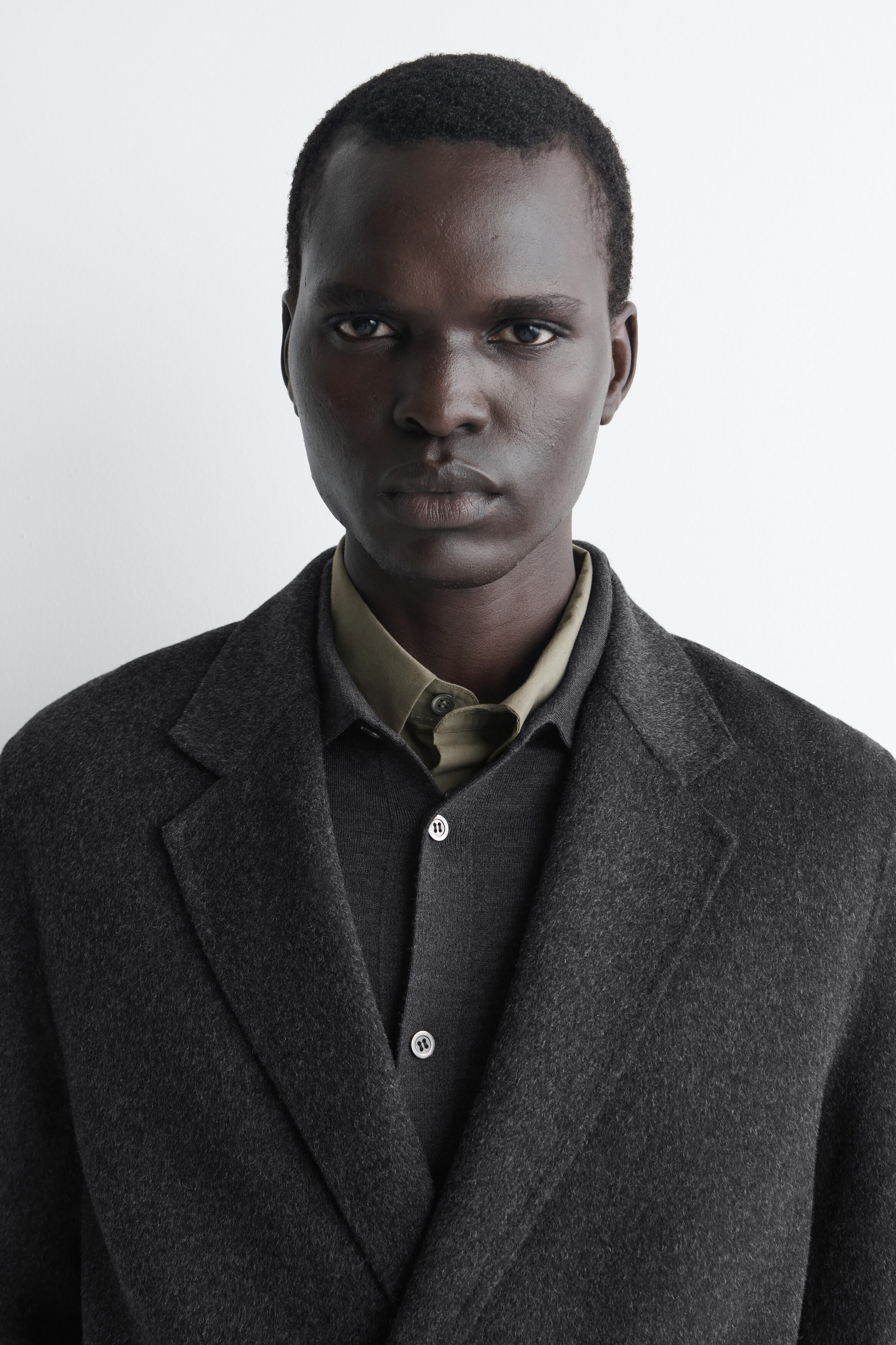A dark grey wool-blend blazer with notched lapels is shown in a close-up front view, layered over a matching dark grey button-up shirt and an olive-green collared shirt. A dark grey wool-blend blazer with notched lapels is shown in a close-up front view, layered over a matching dark grey button-up shirt and an olive-green collared shirt.