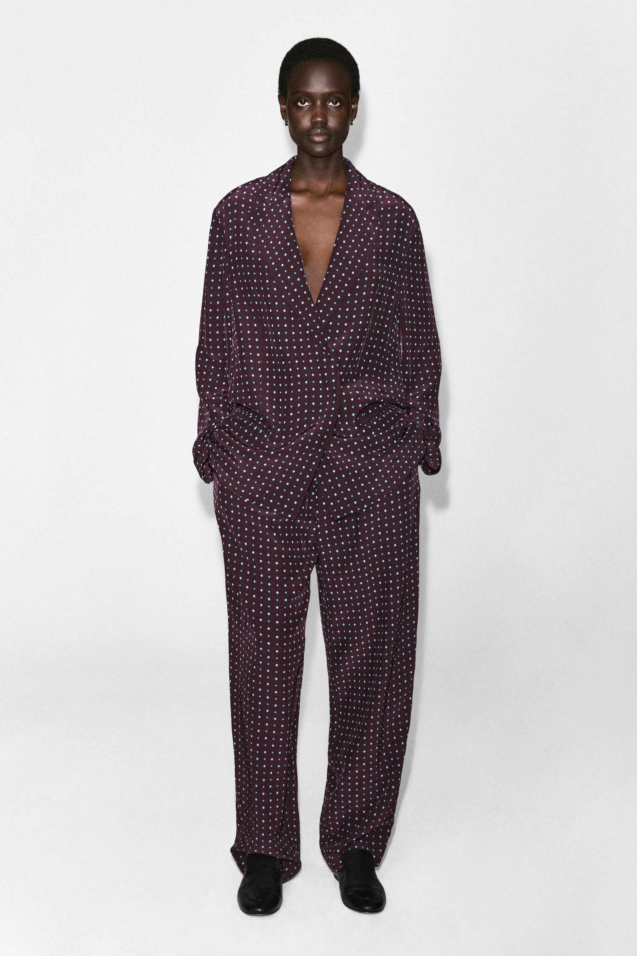 Le pantalon droit à motif losanges est présenté de face dans une coupe décontractée à jambe droite. Il est confectionné dans un tissu bordeaux foncé avec un petit motif répétitif de losanges bleu clair et tombe souplement sur des chaussures noires, en coordination avec un blazer cache-cœur assorti.