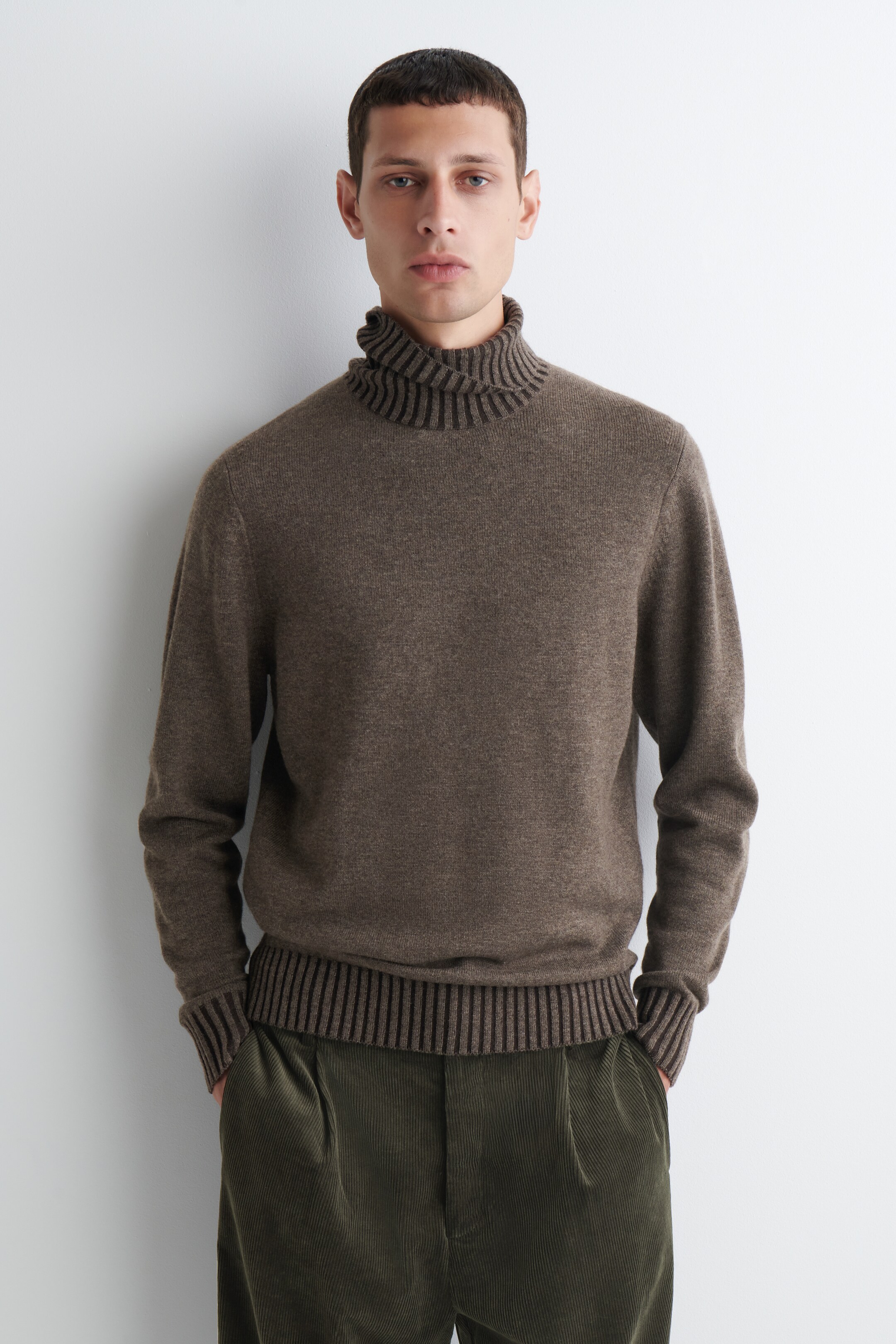 View larger image: The brown turtleneck sweater is featuring a high, folded collar with prominent horizontal dark brown stripes integrated into its ribbed texture, and a plain knit body with ribbed cuffs and a ribbed hem. The sweater is being shown from the front, with its hem resting just above green corduroy pants, showcasing its regular fit.