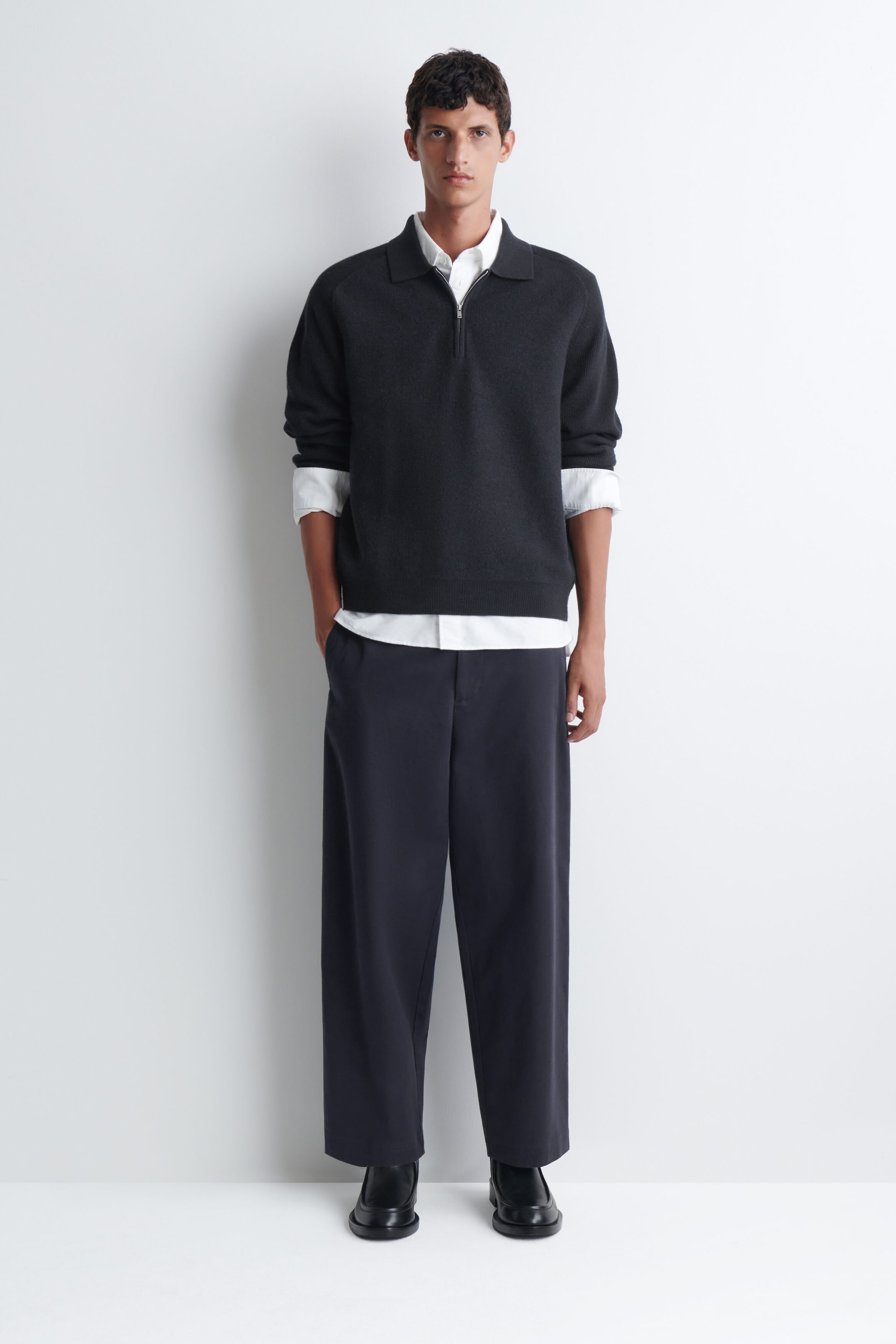 The model is wearing a dark grey boiled-wool zip-up sweater featuring a polo collar and half-zip closure. The sweater is layered over a white collared shirt, with the hem and rolled cuffs visible, and styled with dark wide-leg trousers. The model is wearing a dark grey boiled-wool zip-up sweater featuring a polo collar and half-zip closure. The sweater is layered over a white collared shirt, with the hem and rolled cuffs visible, and styled with dark wide-leg trousers.