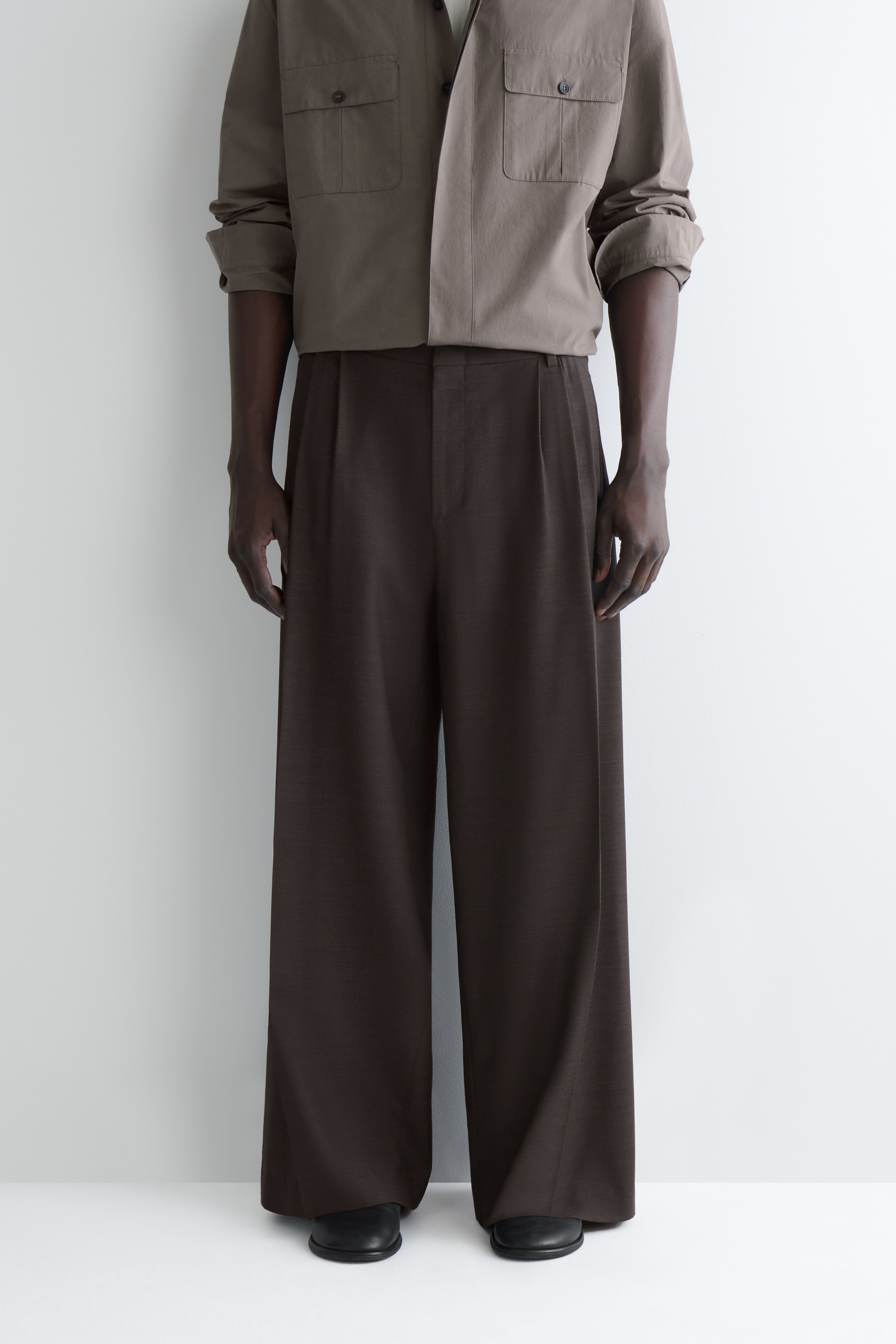 Grösseres Bild ansehen: Dunkelbraune Hosen mit weitem Bein werden von vorne präsentiert, mit Falten an der Taille und einem Schnitt in voller Länge, der über die Schuhe fällt.