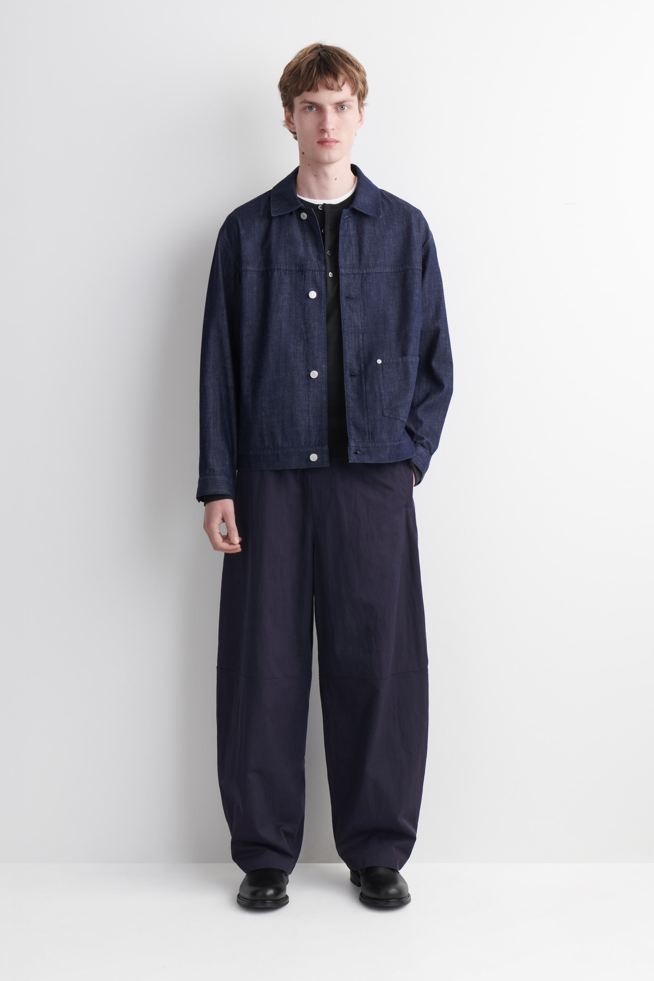 Ver imagen más grande: Una chaqueta de mezclilla azul oscuro presenta un cuello en punta, botones frontales blancos y dos bolsillos delanteros de parche, usada sobre una camiseta Henley negra. Los pantalones de pierna ancha en azul oscuro están diseñados con un ajuste relajado y detalles de costura en las rodillas.