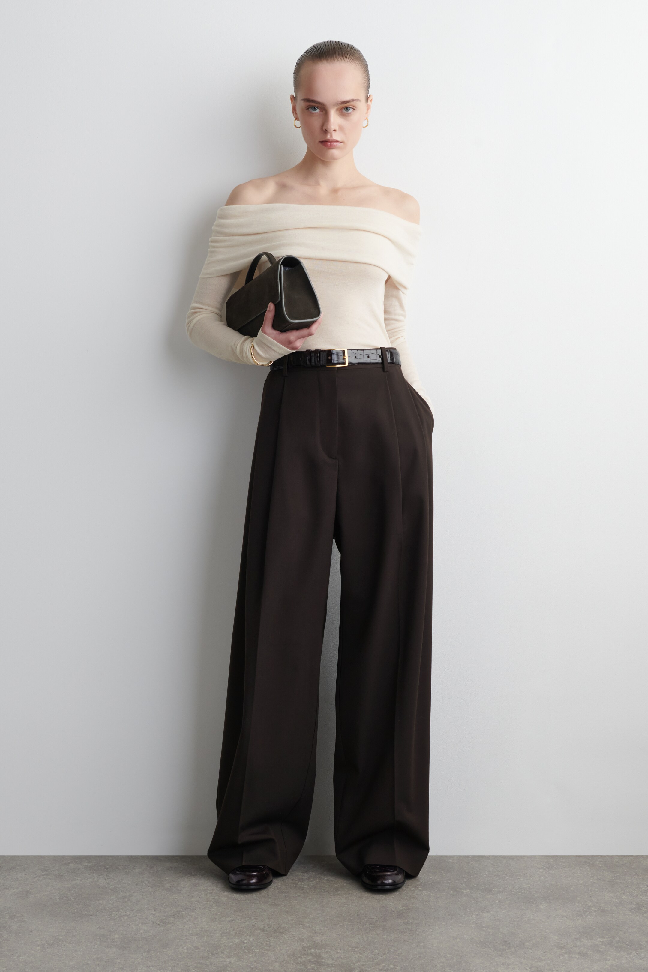 Agrandir l'image: Le produit est un haut en maille fine de couleur blanc cassé, à manches longues, qui se porte sur les épaules et est présenté de face. Il est stylé avec un pantalon large à taille haute brun foncé, avec des plis à l'avant et ceinturé par une ceinture noire avec une boucle dorée.