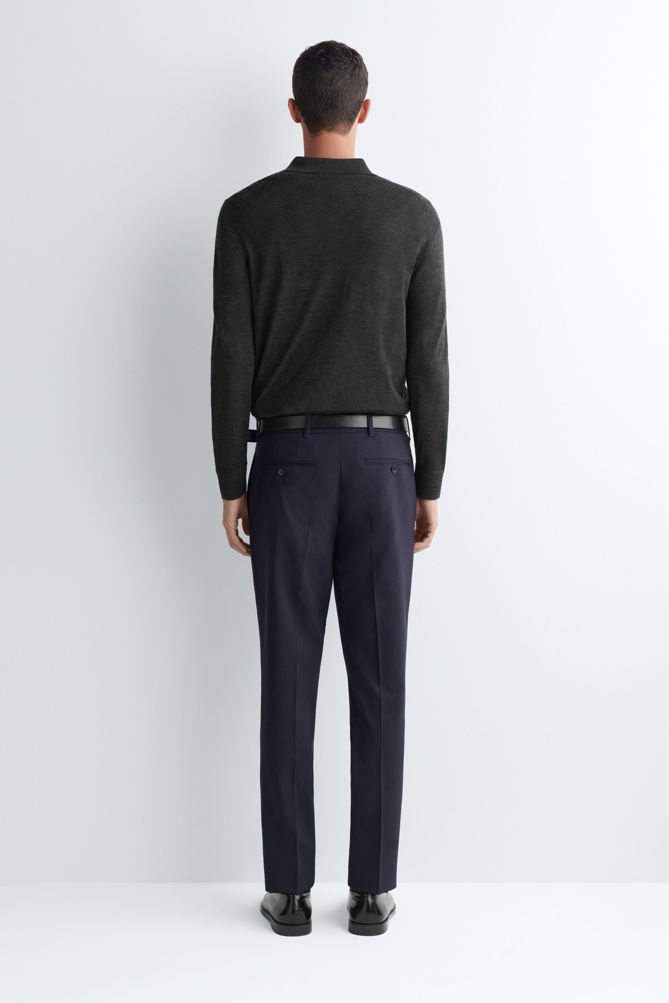 Un pantalon droit slim en laine bleu marine foncé est porté par le mannequin, vu de dos. Le pantalon présente des plis marqués au fer sur le devant de chaque jambe et des poches passepoilées boutonnées, porté avec une ceinture en cuir noir et un polo à manches longues gris foncé rentré dans le pantalon. Un pantalon droit slim en laine bleu marine foncé est porté par le mannequin, vu de dos. Le pantalon présente des plis marqués au fer sur le devant de chaque jambe et des poches passepoilées boutonnées, porté avec une ceinture en cuir noir et un polo à manches longues gris foncé rentré dans le pantalon.