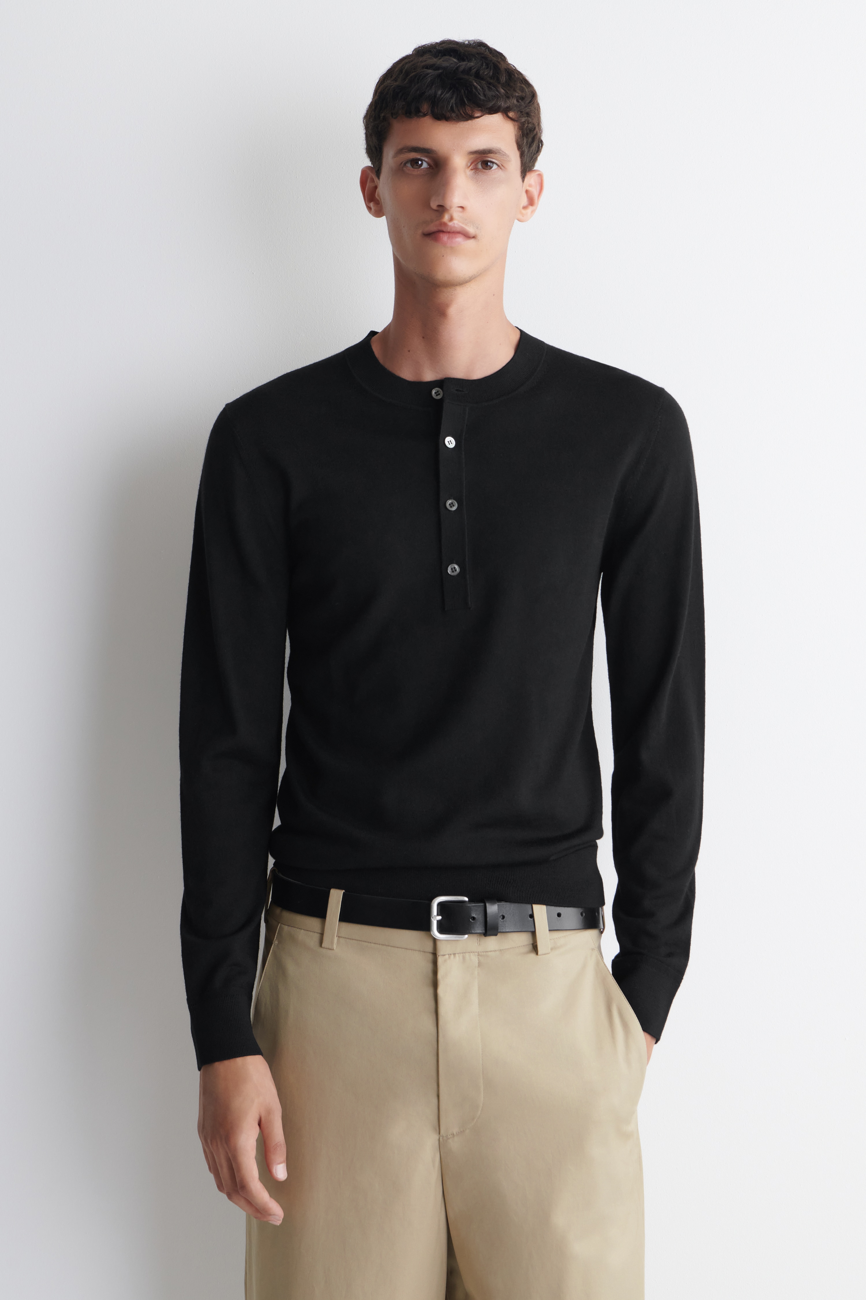 Czarny longsleeve typu Henley jest pokazany z przodu. Wyróżnia się drobną dzianinową fakturą, okrągłym dekoltem z rozpinanym zapięciem na guziki oraz pięcioma jasnymi guzikami. Bluzka ma prążkowane mankiety i prążkowany dół.