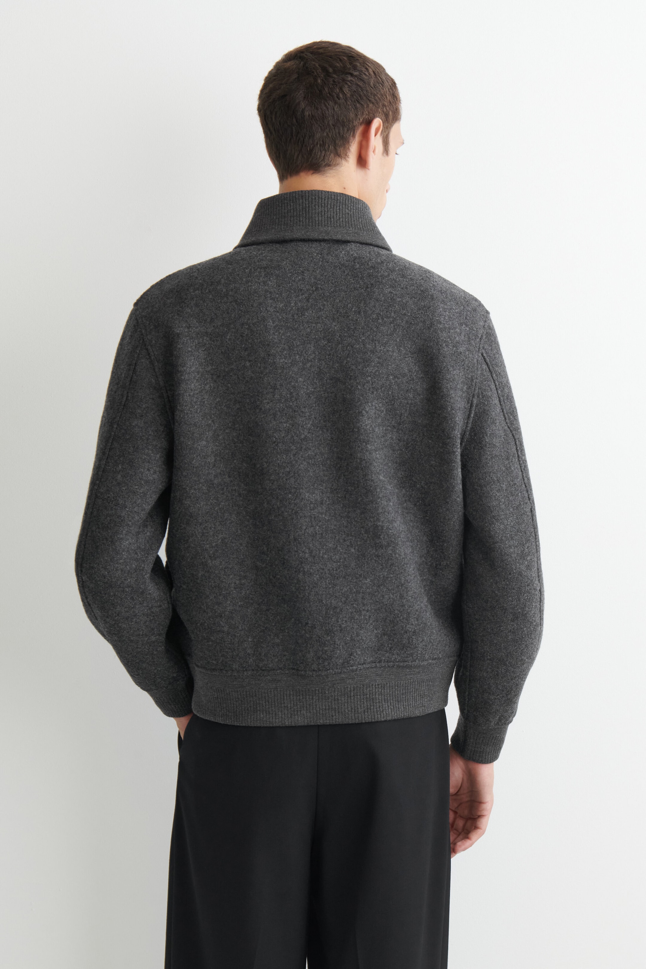 The boiled wool shawl-collar jacket is shown from the back, highlighting its structured fit and textured dark gray fabric. It features a ribbed collar, ribbed cuffs, and a ribbed hem, and is paired with black trousers. The boiled wool shawl-collar jacket is shown from the back, highlighting its structured fit and textured dark gray fabric. It features a ribbed collar, ribbed cuffs, and a ribbed hem, and is paired with black trousers.