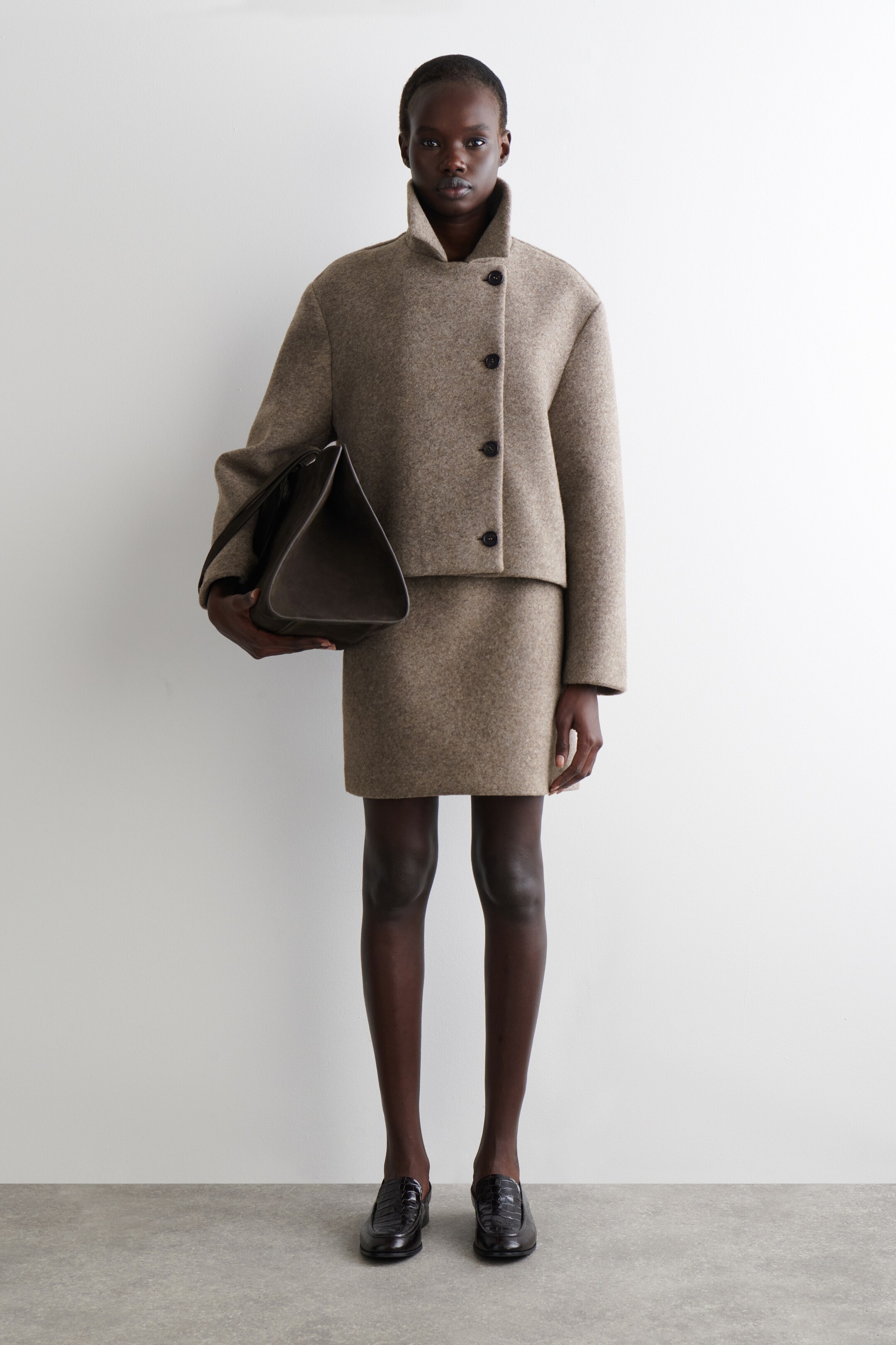 Visualizza immagine più grande: Una giacca beige testurizzata, corta, con una chiusura asimmetrica a bottoni scuri e un colletto alto viene presentata frontalmente, abbinata a una mini gonna coordinata. Una borsa a mano scura e strutturata è tenuta in una mano, e mocassini scuri e testurizzati sono visibili ai piedi.