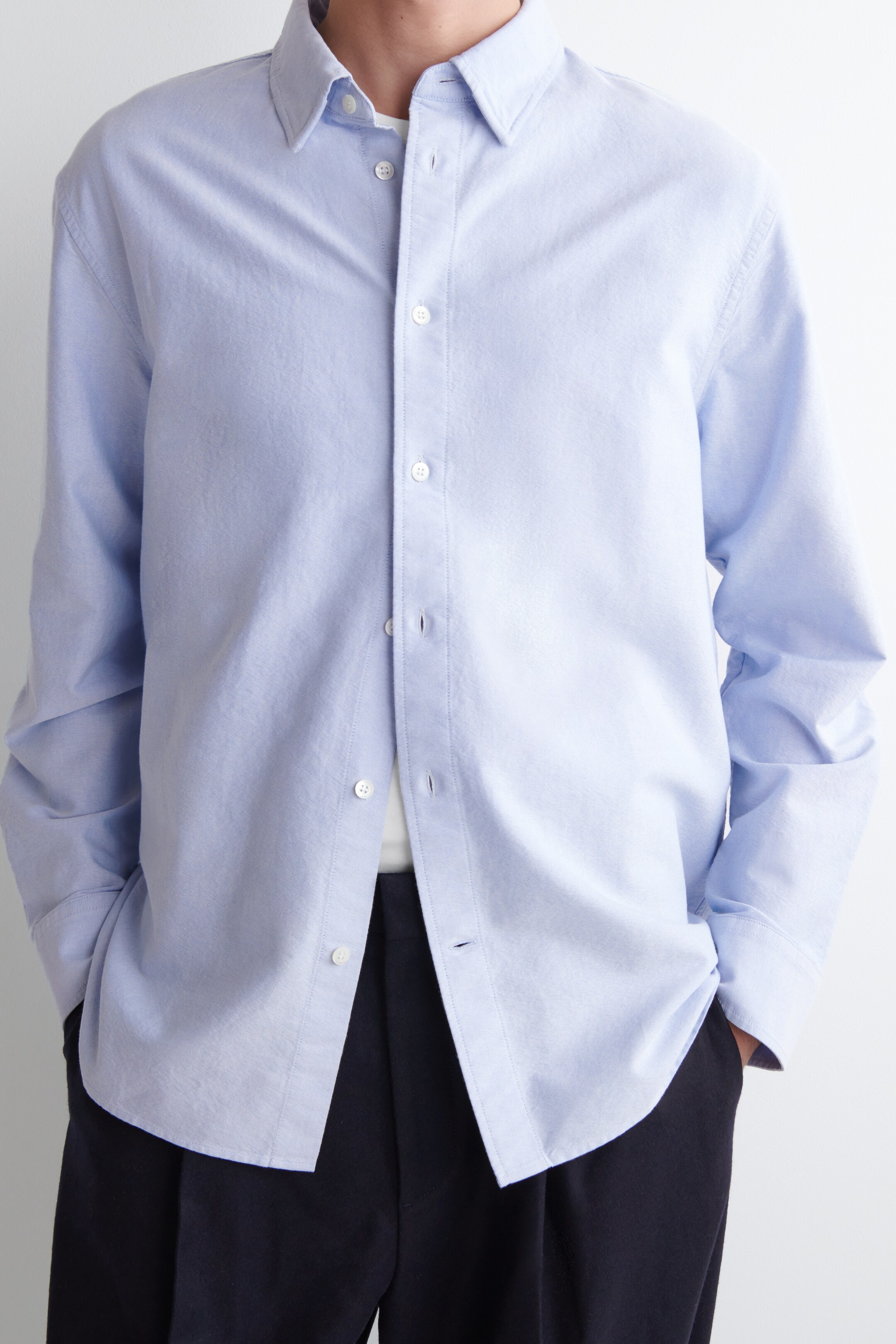 Ver imagen más grande: Una camisa azul claro de botones, hecha de un tejido sutilmente texturizado, se muestra desde el frente. Presenta un cuello de botones, una tapeta completa con botones blancos y mangas largas, ofreciendo un ajuste relajado mientras se lleva sin meter sobre pantalones oscuros y revelando una camiseta blanca debajo.