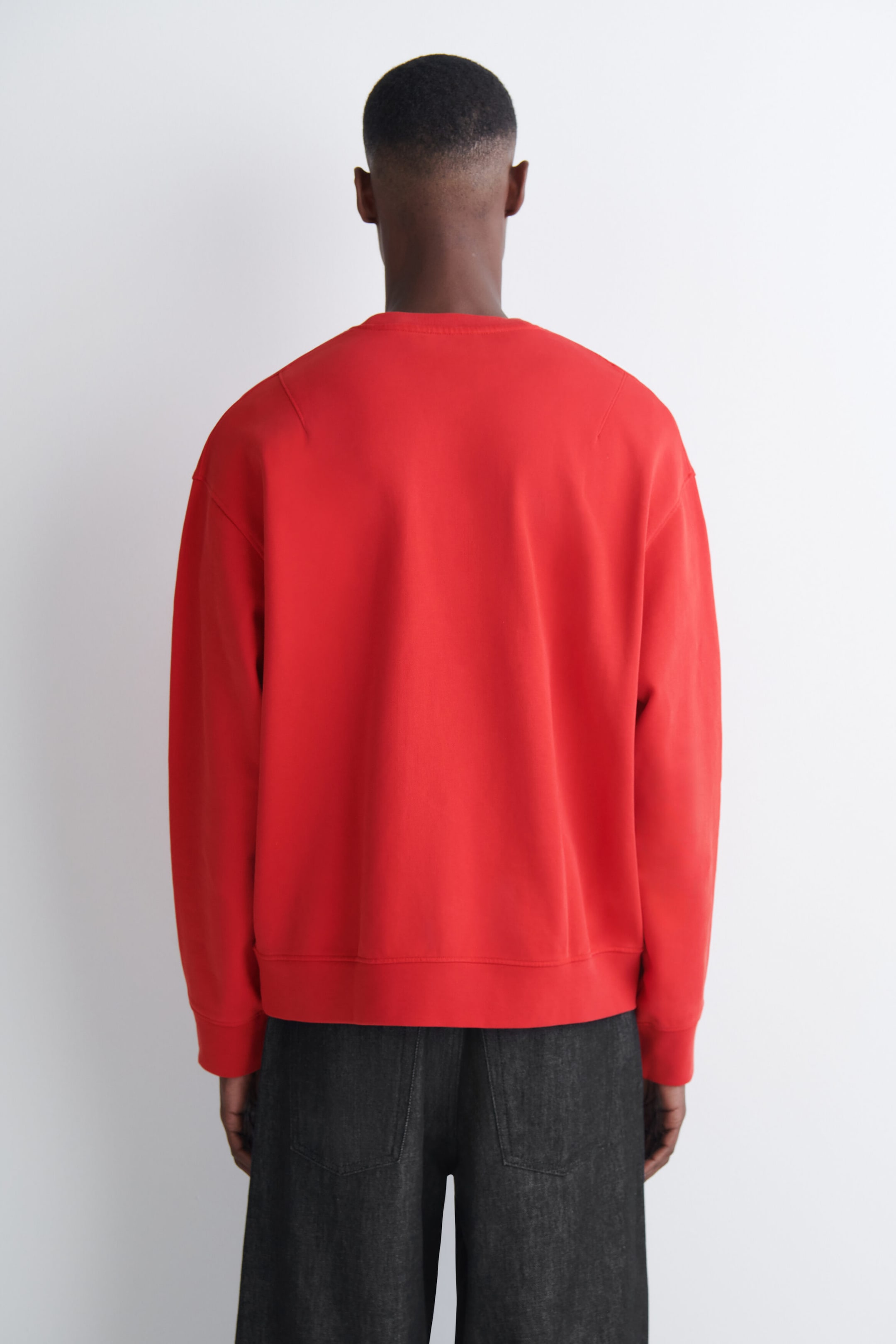 A red, relaxed-fit crew-neck sweatshirt is being worn by the model, who is facing away from the camera. The sweatshirt, featuring ribbed trims at the hem and cuffs, is being paired with dark denim trousers. A red, relaxed-fit crew-neck sweatshirt is being worn by the model, who is facing away from the camera. The sweatshirt, featuring ribbed trims at the hem and cuffs, is being paired with dark denim trousers.