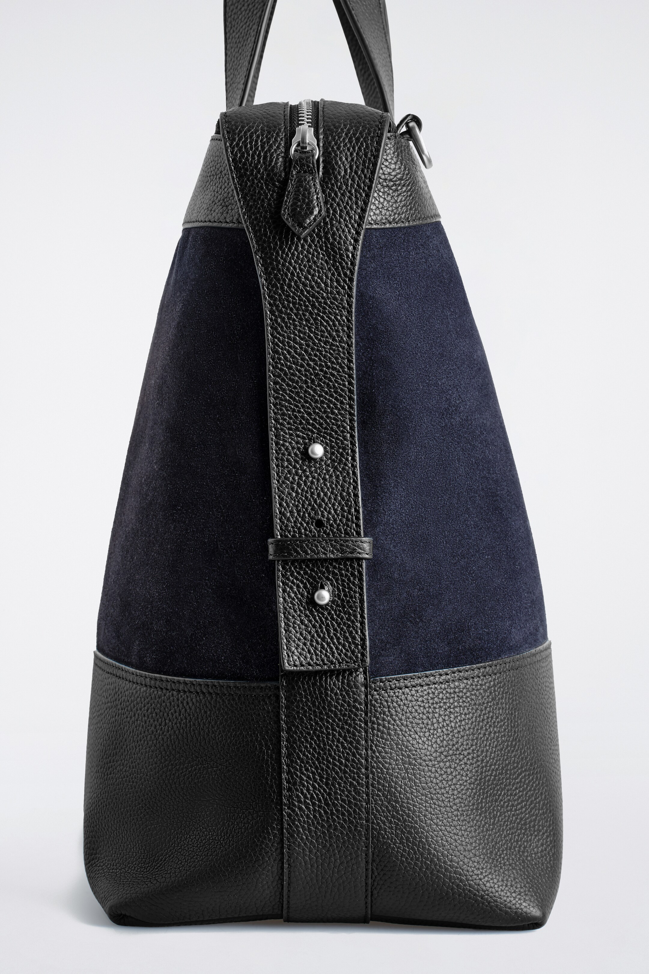 Visualizza immagine più grande: Una borsa strutturata è in piedi, vista da un angolo inferiore, mostrando il suo fronte e il lato. Presenta un corpo principale in camoscio blu scuro, arricchito da pannelli in pelle nera a grana alla base e in cima. Una cinghia verticale in pelle nera a grana corre lungo il fronte, fissata da due rivetti color argento.