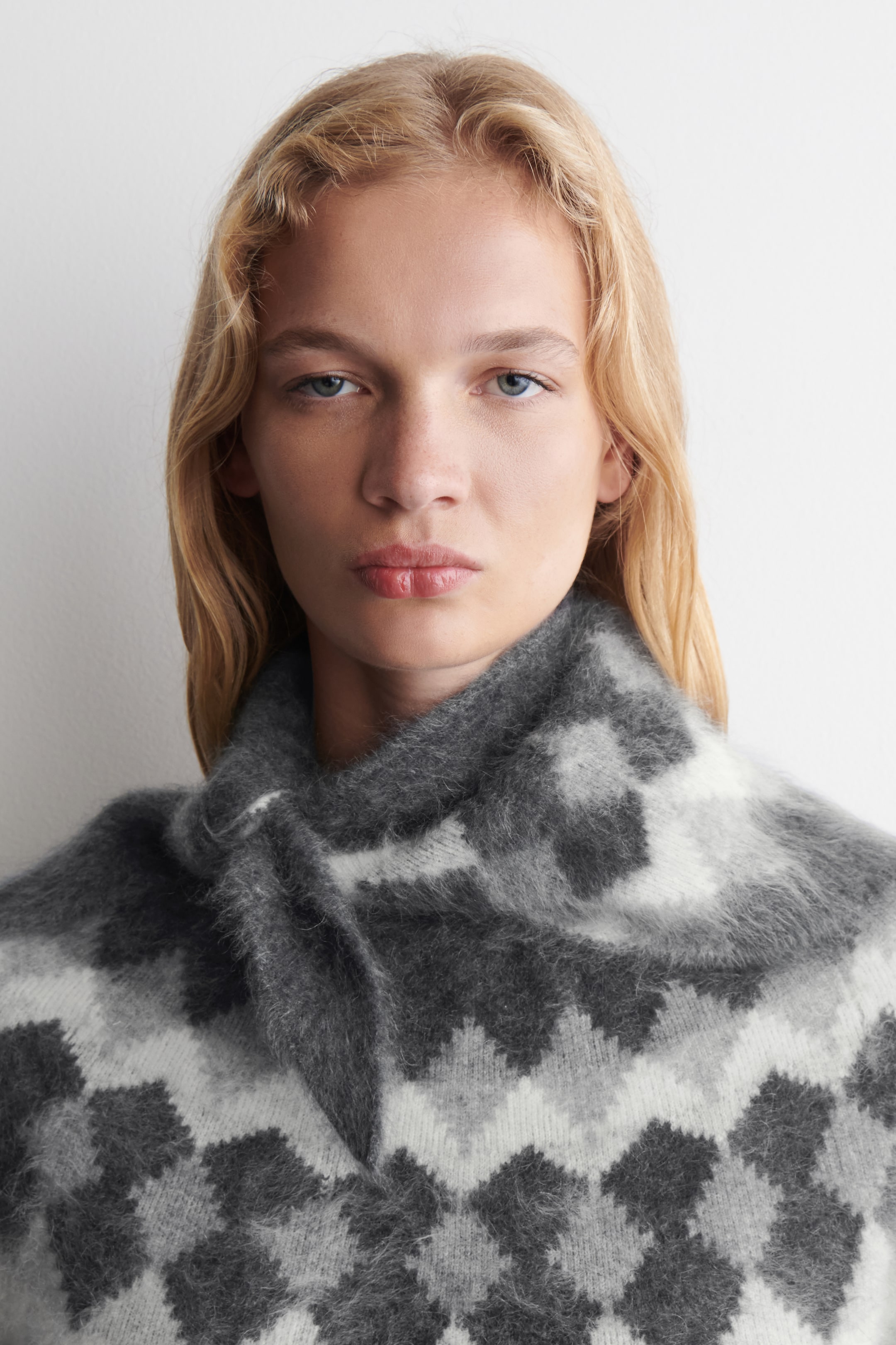 Ver imagen más grande: Un jersey de punto peludo presenta un patrón geométrico en tonos grises y blancos, con formas de diamante y triángulo. Su cuello alto se drapea suavemente alrededor del cuello y los hombros del usuario.