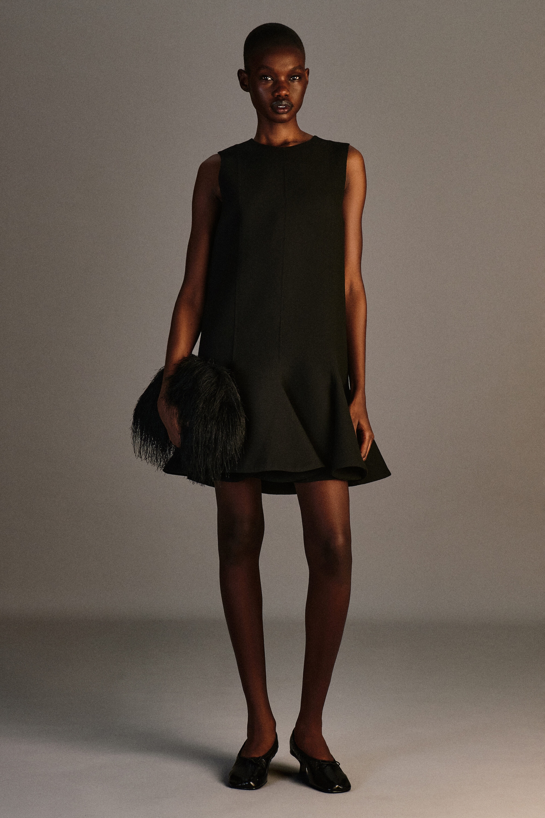 A black wool-blend twill peplum mini dress is shown from the front, featuring a sleeveless design and a high round neckline. The structured, straight-cut bodice flares out at the hem into a dramatic, ruffled peplum. A black wool-blend twill peplum mini dress is shown from the front, featuring a sleeveless design and a high round neckline. The structured, straight-cut bodice flares out at the hem into a dramatic, ruffled peplum.