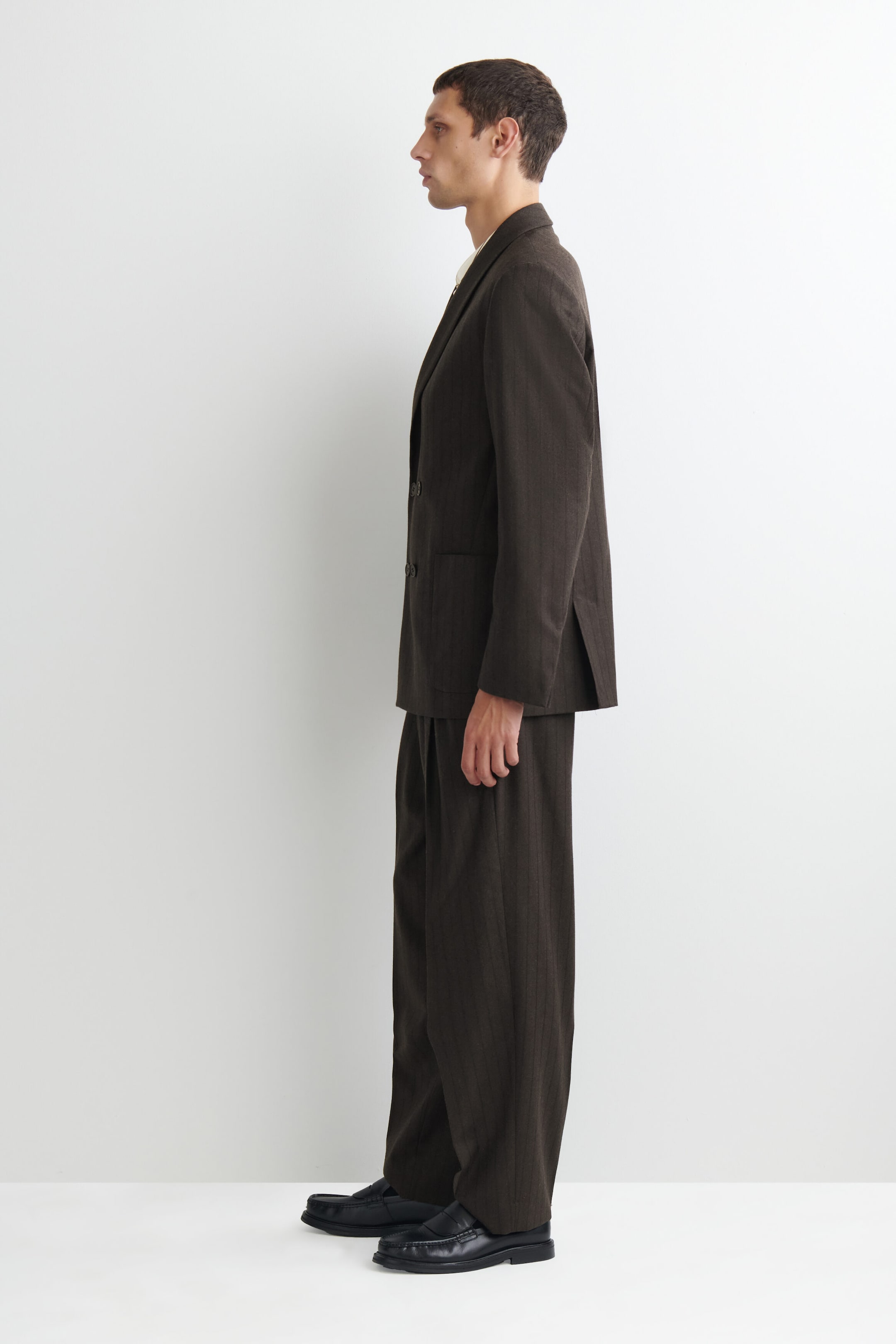 A model is standing in profile, facing left, wearing a dark brown, subtly pinstriped double-breasted blazer paired with matching wide-leg trousers. The relaxed-fit blazer features a split hem at the back and is styled with black leather loafers. A model is standing in profile, facing left, wearing a dark brown, subtly pinstriped double-breasted blazer paired with matching wide-leg trousers. The relaxed-fit blazer features a split hem at the back and is styled with black leather loafers.