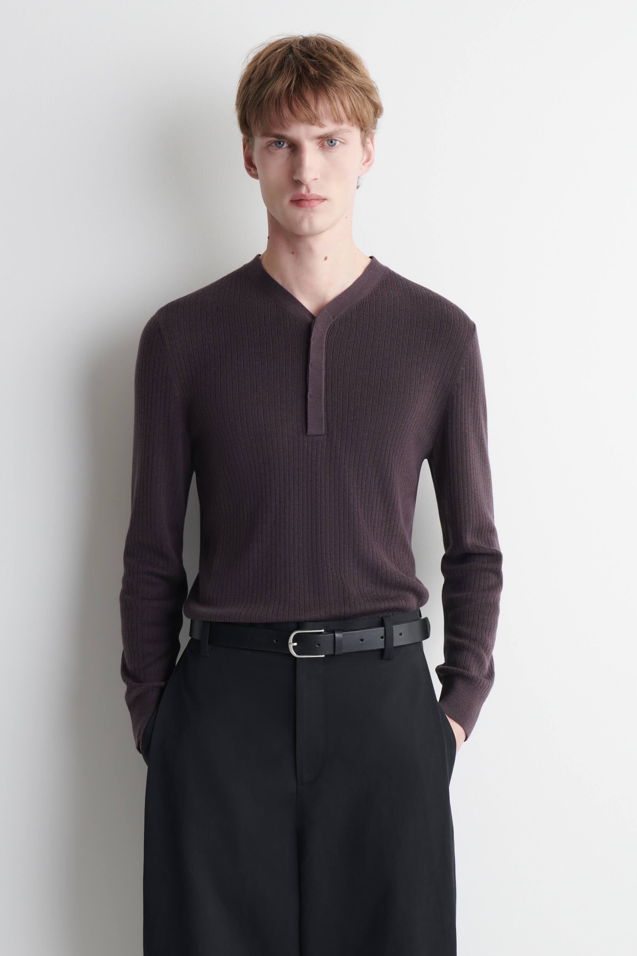 View larger image: The model is wearing a dark plum ribbed knit Henley shirt. It features a ribbed texture, a V-neckline, and a single-button placket, and is styled with black trousers and a black belt.
