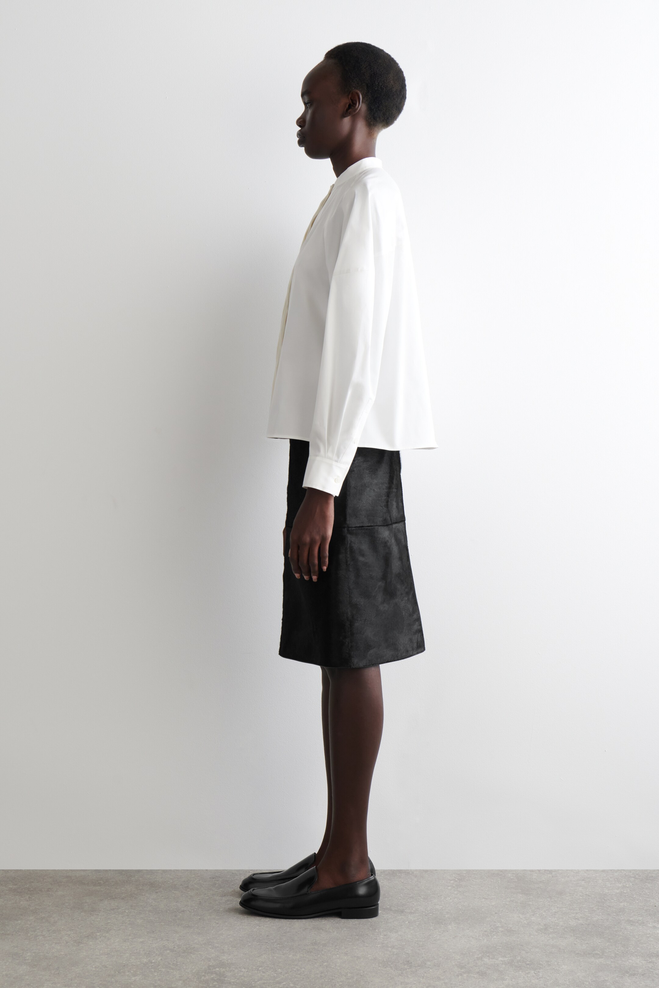 View larger image: A white, loose-fitting shirt with a band collar and long buttoned cuffs is being presented in profile, featuring a light-colored tie detail at the neckline, and is styled with a black, textured, knee-length A-line skirt showing a vertical side seam. Black leather loafers with a low heel are completing the ensemble.