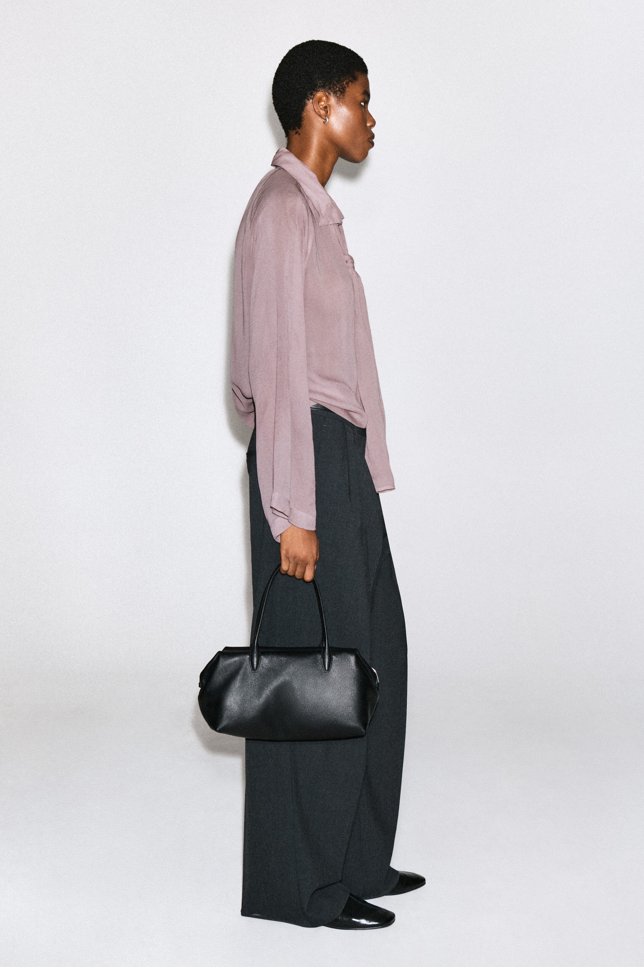 Un haut fluide à manches longues, dans une teinte mauve poudré, est porté par le mannequin. Il présente un col drapé et une silhouette décontractée. Le haut est associé à un pantalon large foncé, tandis que le mannequin tient un sac à main en cuir noir.
