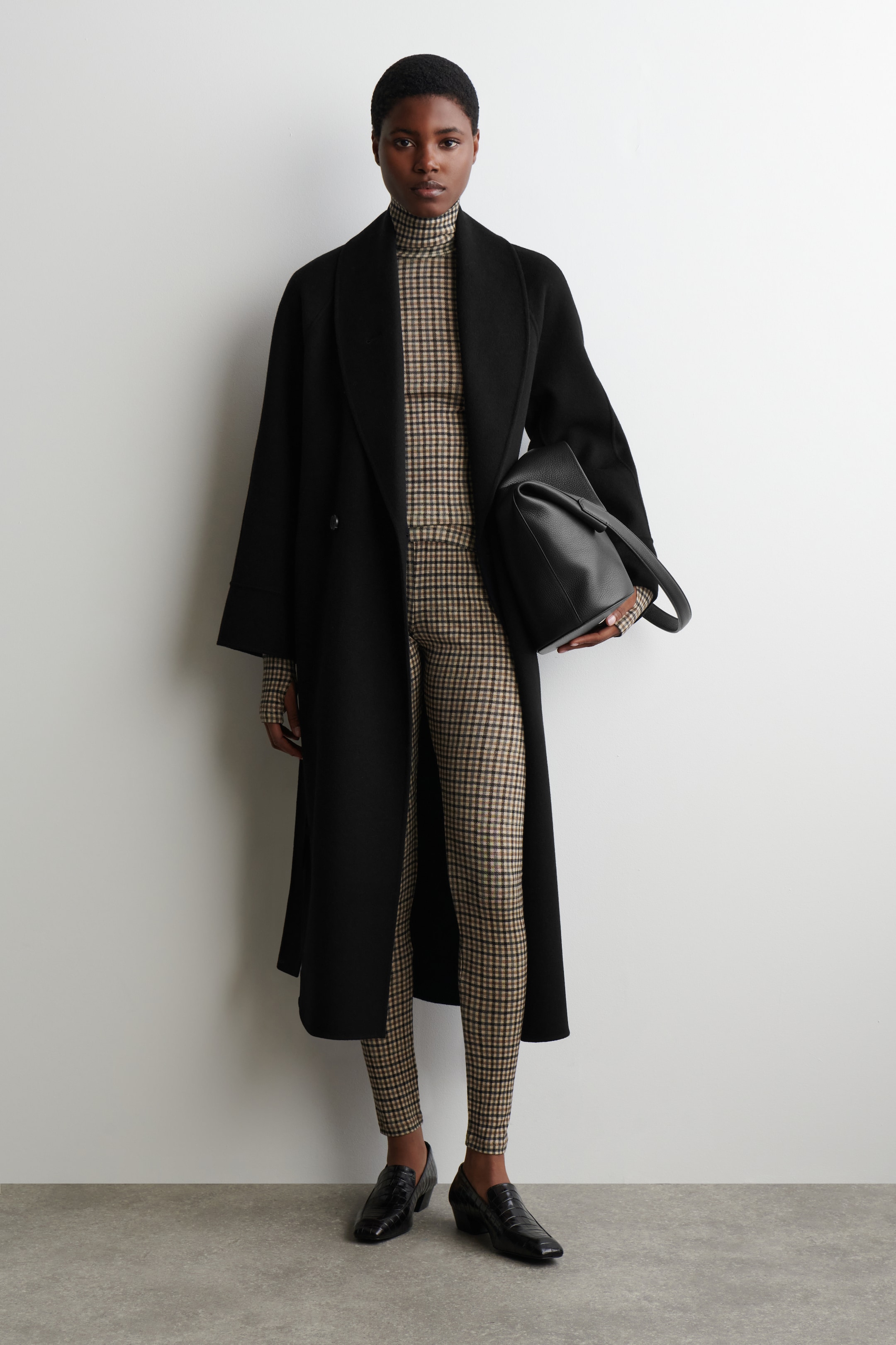 Visualizza immagine più grande: Leggings attillati con un motivo a quadri beige e marrone scuro sono indossati dalla modella. Sono abbinati a un dolcevita coordinato e stratificati sotto un lungo cappotto nero.