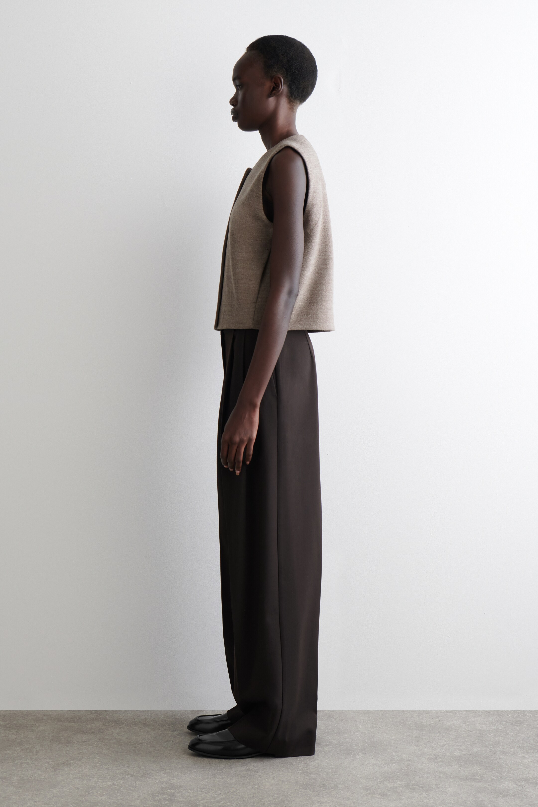 Agrandir l'image: Un gilet texturé, chiné beige est porté par un mannequin orienté vers la gauche, avec un col rond, une fermeture à boutons à l'avant, des coutures frontales angulaires et une longueur courte.