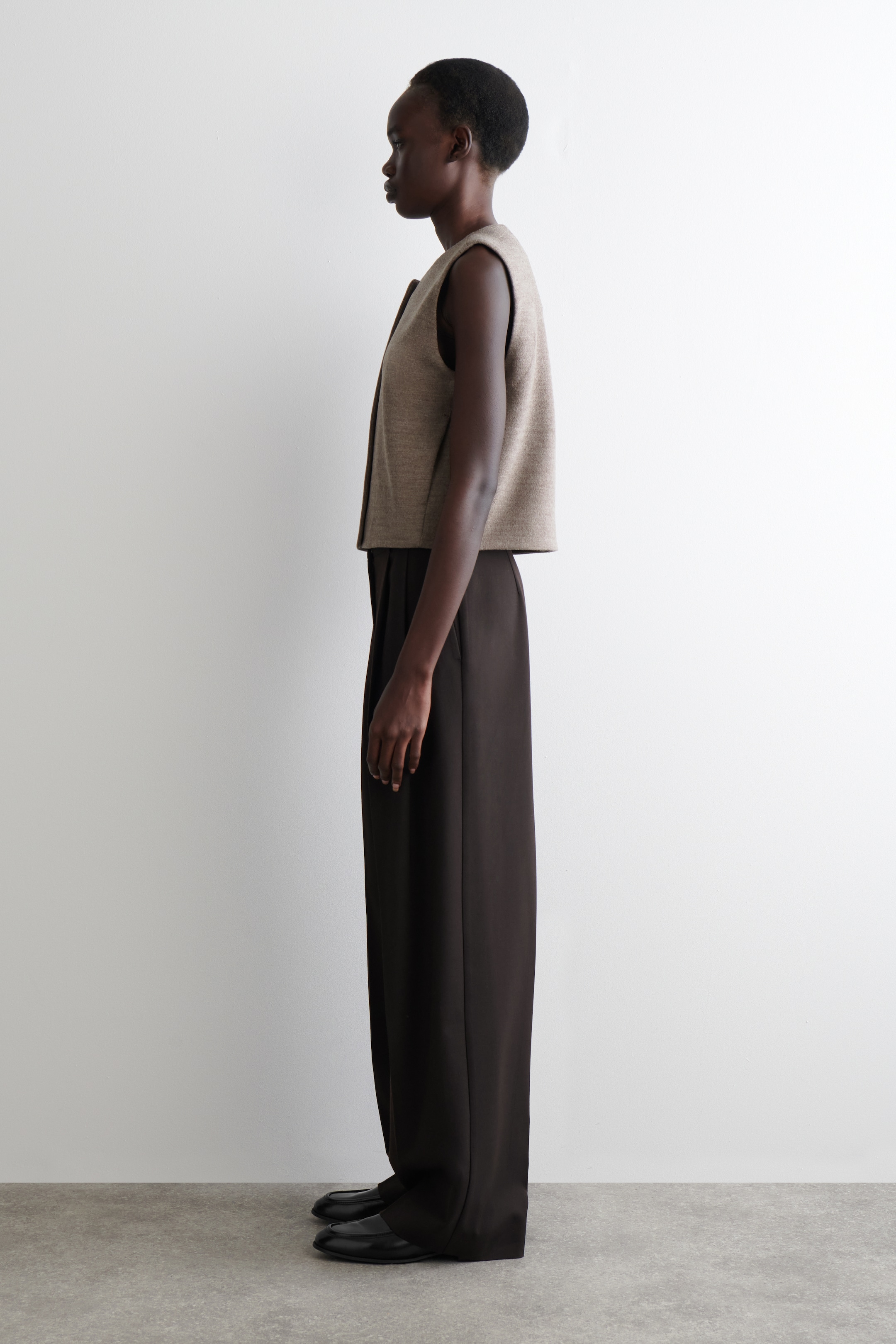 View larger image: A textured, heathered beige waistcoat is being worn by a model facing left, featuring a rounded neckline, a button-front closure, angular front seams, and a cropped length.