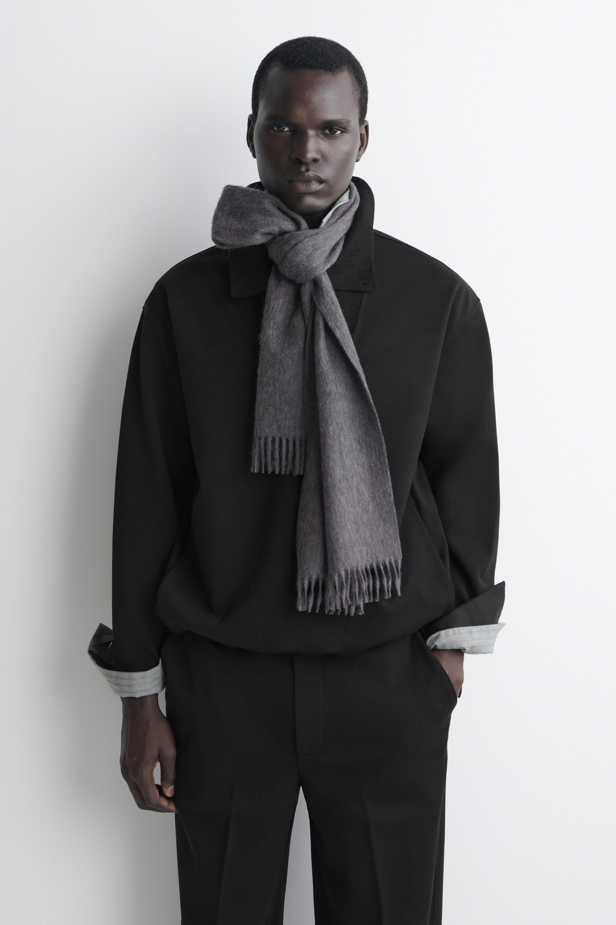 A grey cashmere scarf is being worn in a front-facing view, looped in a knot around the neck over a black jacket. The scarf features a soft, textured weave with fringed trim on both ends hanging down the torso. A grey cashmere scarf is being worn in a front-facing view, looped in a knot around the neck over a black jacket. The scarf features a soft, textured weave with fringed trim on both ends hanging down the torso.