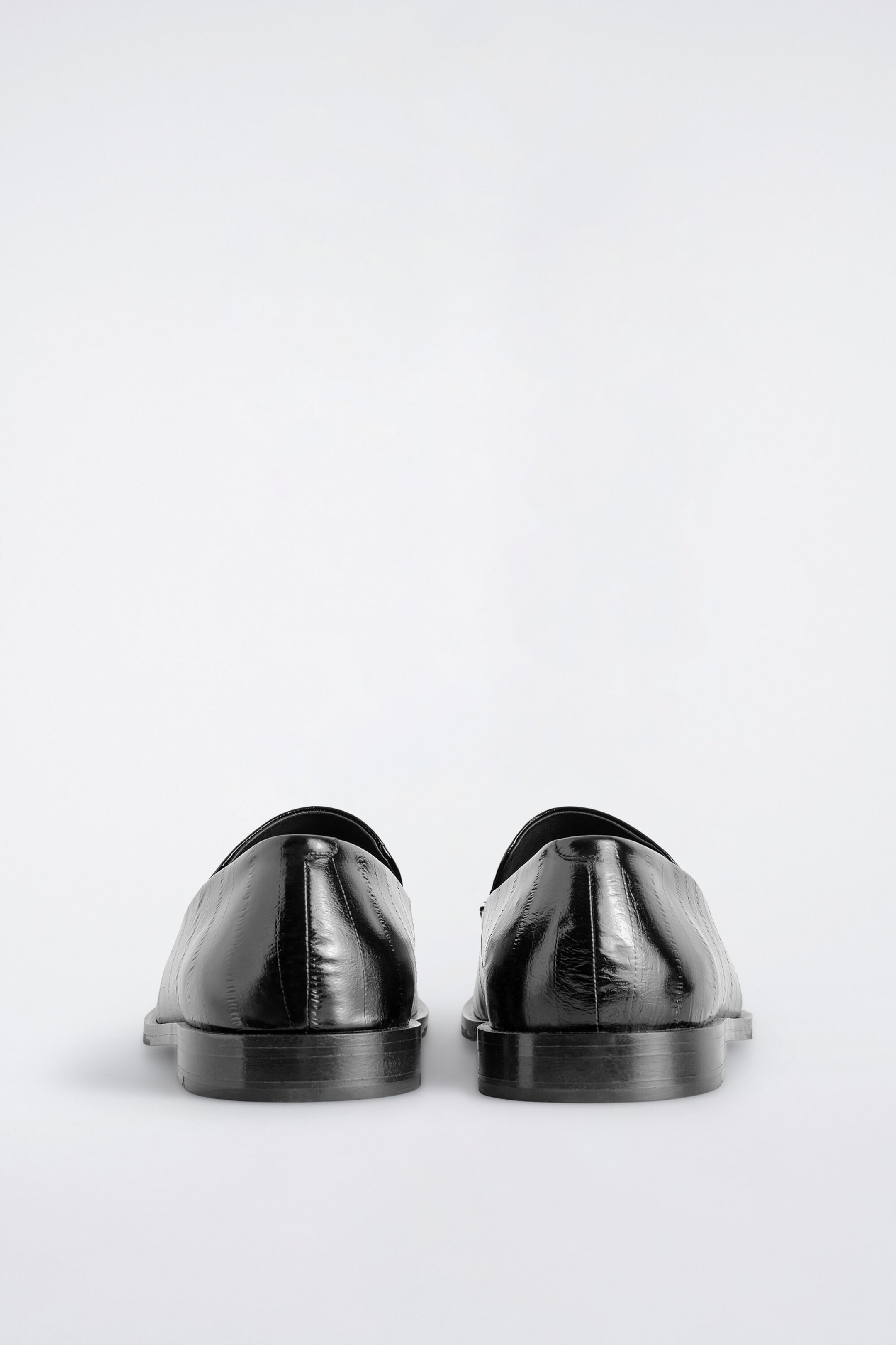 A pair of black polished leather loafers stands side by side, viewed from the back to show the heel structure and low block heels. The glossy leather features a subtle, vertically oriented eel-effect embossed texture. A pair of black polished leather loafers stands side by side, viewed from the back to show the heel structure and low block heels. The glossy leather features a subtle, vertically oriented eel-effect embossed texture.