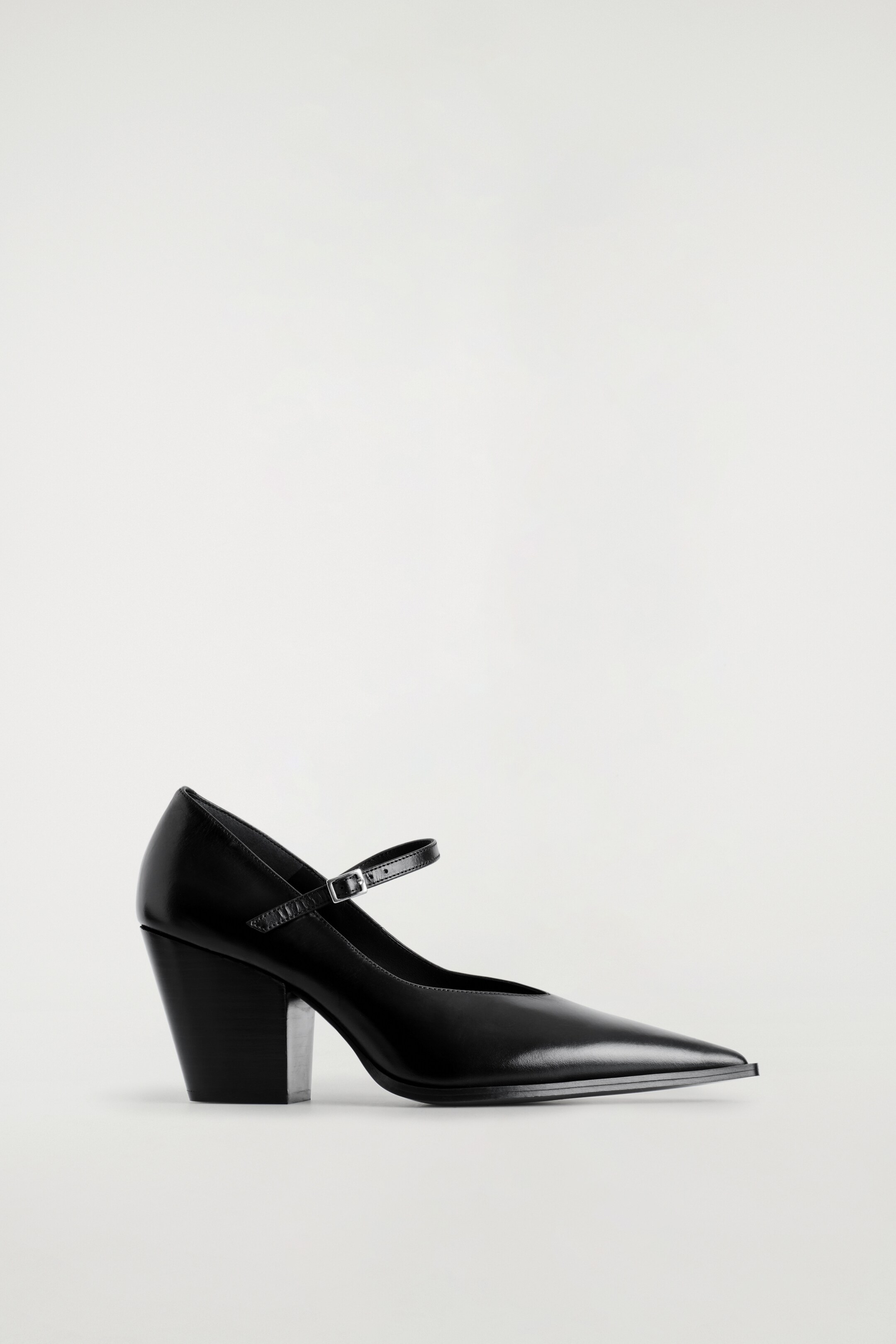 Agrandir l'image: Une chaussure Mary Jane à talon en cuir noir est exposée, inclinée vers la gauche, montrant son côté droit. Elle présente un bout pointu, une seule bride avec une boucle argentée traversant le cou-de-pied, et un talon bloc épais avec une surface intérieure réfléchissante.