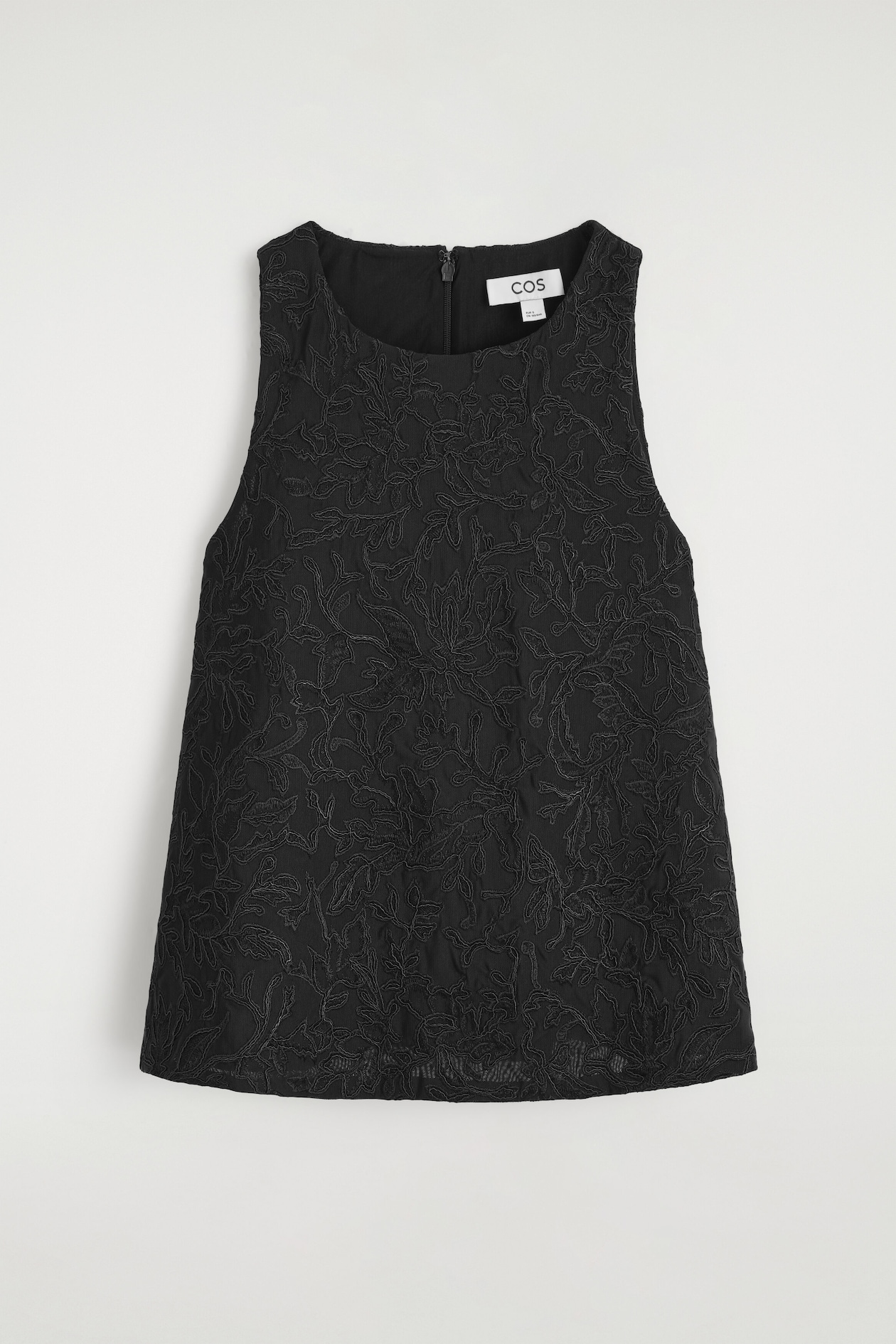 Un haut noir sans manches est présenté de face, mettant en valeur une broderie florale complexe sur toute la surface, sur un fond en maille transparente. Le haut présente une silhouette trapèze décontractée avec un col rond montant et une fermeture éclair visible à l’arrière du col.